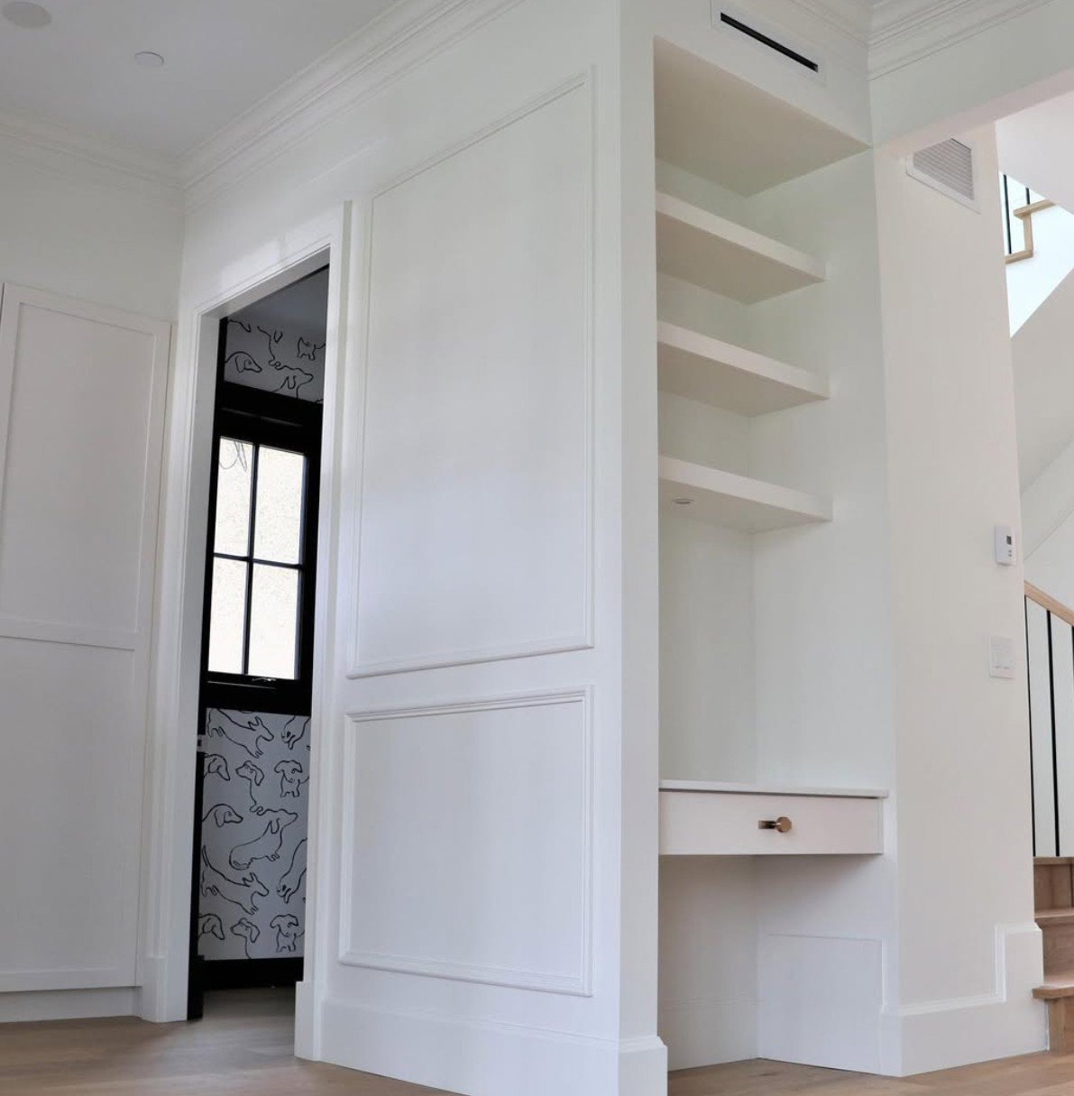 Interior view of a white wall with built-in shelves, a small drawer, and a staircase with black railing. A window with a framed design is visible in the background.