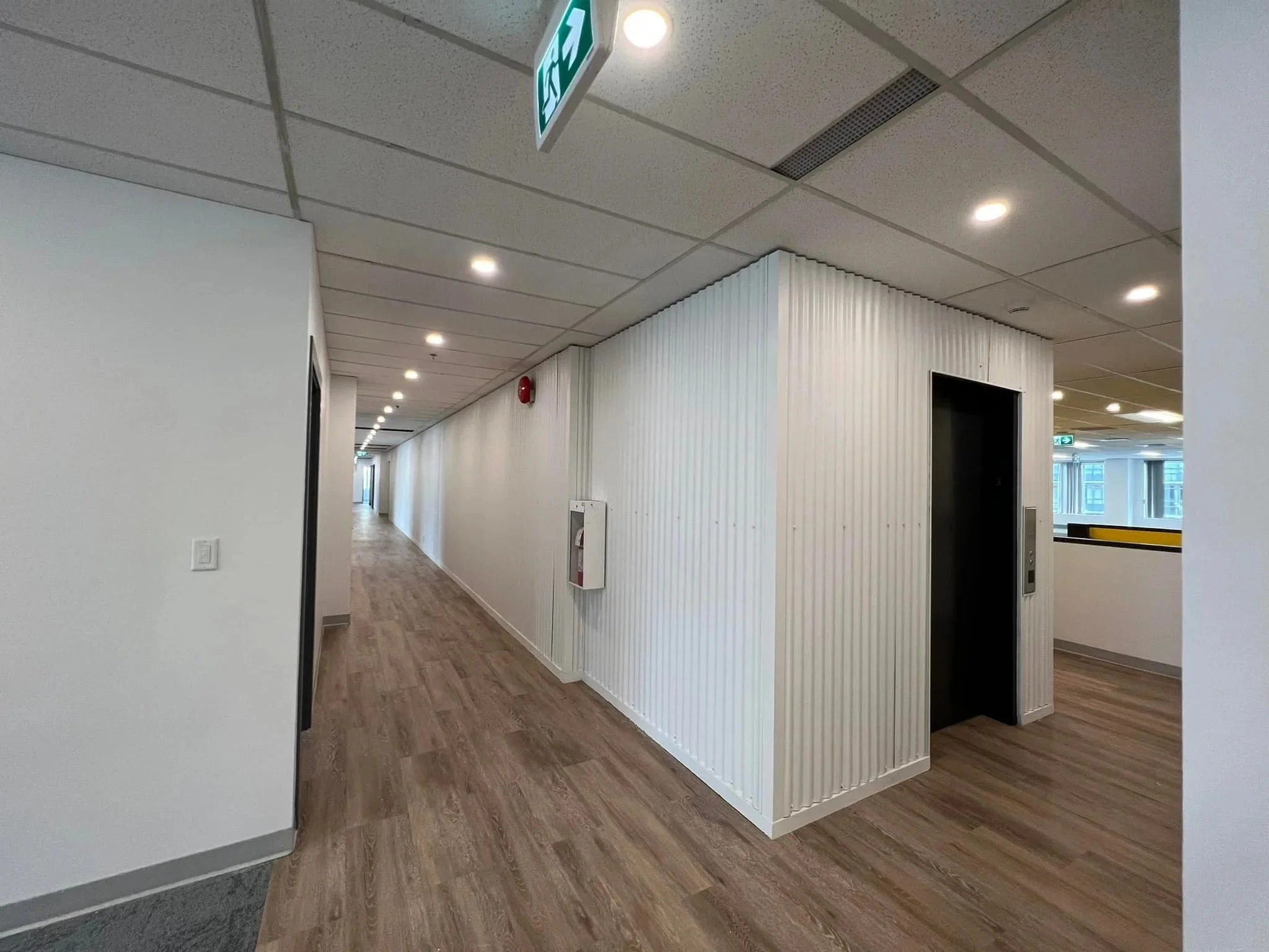 Empty office corridor with wood flooring, white walls, and ceiling lights; elevator door visible on the right, and emergency exit signs hanging from the ceiling.