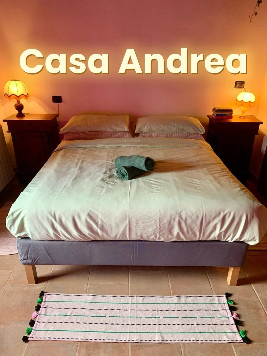 A cozy bedroom with a bed, two bedside tables with lamps, a stack of books, and a colorful sliding rug on a tiled floor. Text overlay: 'Casa Andrea'.