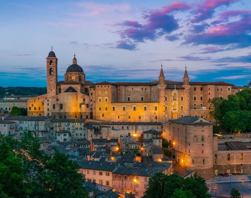 Sunset view of a historic European castle with illuminated stone walls and surrounding buildings, under a colorful evening sky with pink and blue clouds.