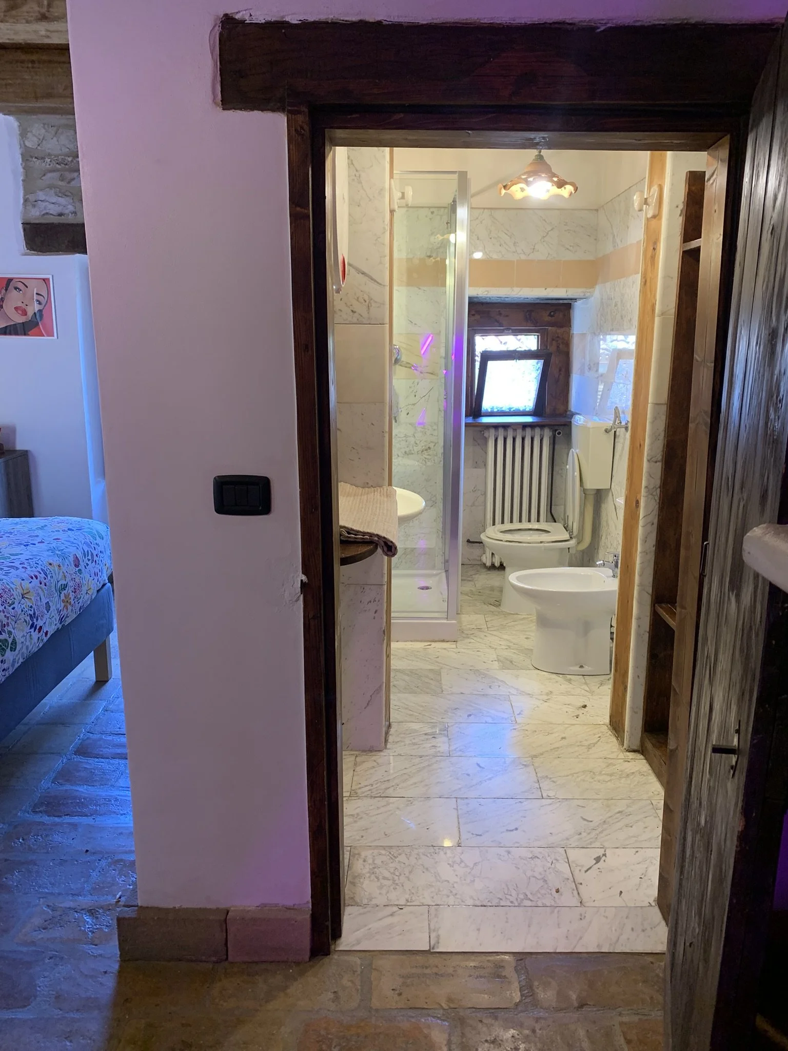 View of a rustic bathroom with marble tiles, a shower enclosure, a toilet, and a small window, viewed through a doorway with a wooden frame.