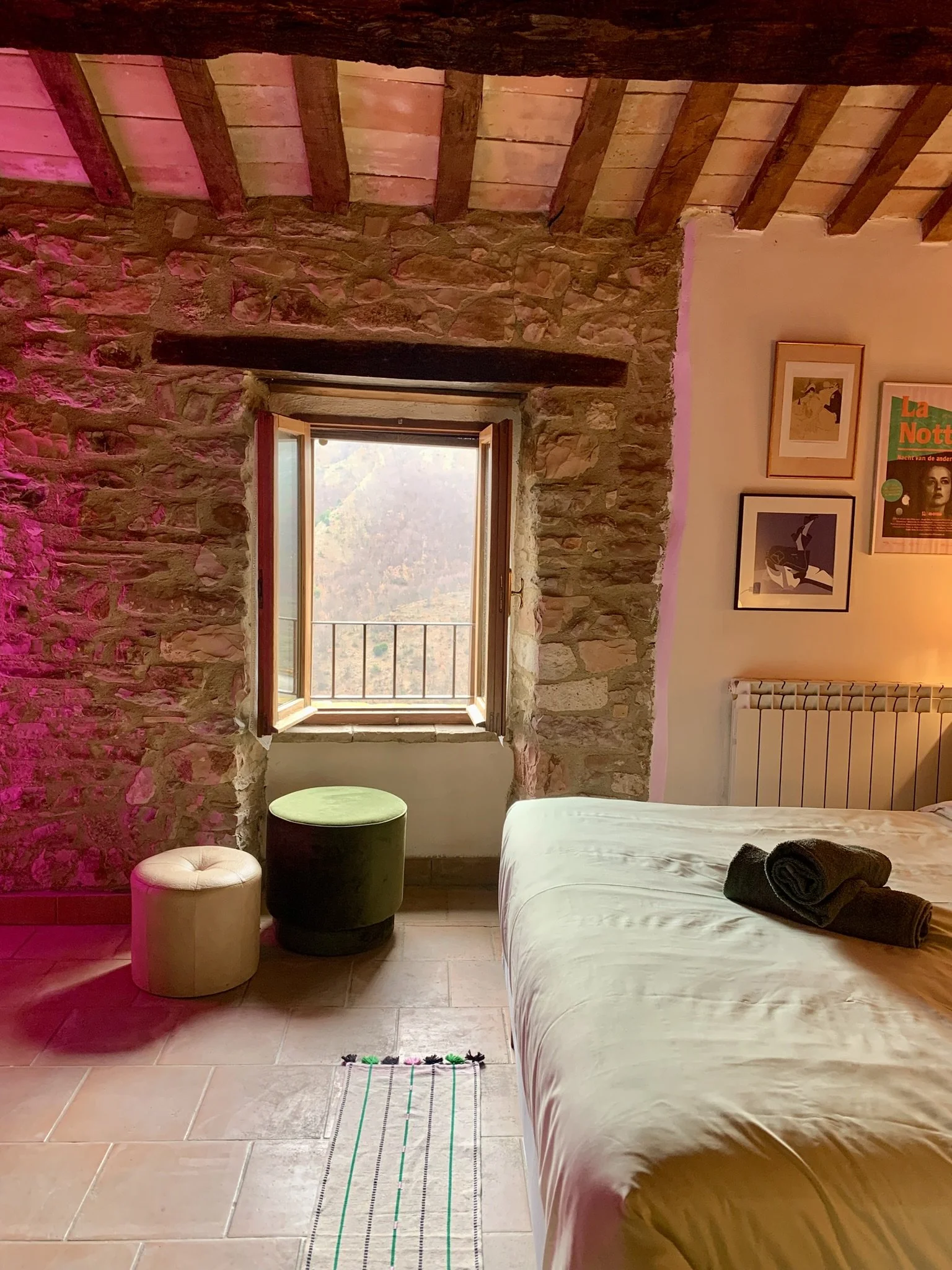 A bedroom with stone and brick walls, a wooden ceiling with beams, a window with a view of trees, a bed with rolled towels, a small round stool, a green cushioned stool, framed artwork on the wall, and tiled flooring.