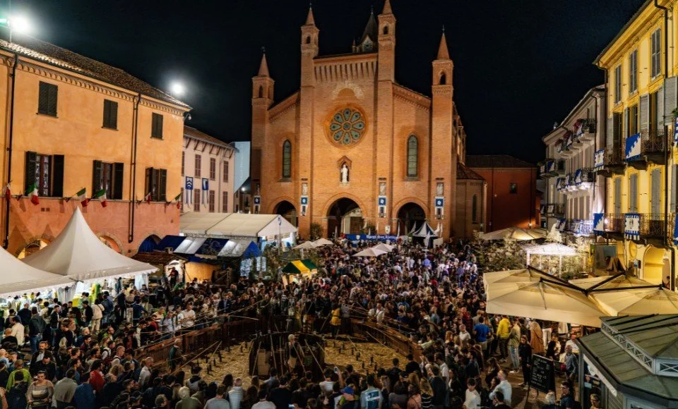 Nighttime scene of a large crowd gathered in front of a historic church with gothic architecture, illuminated by city lights in a European town square, with tents and market stalls.