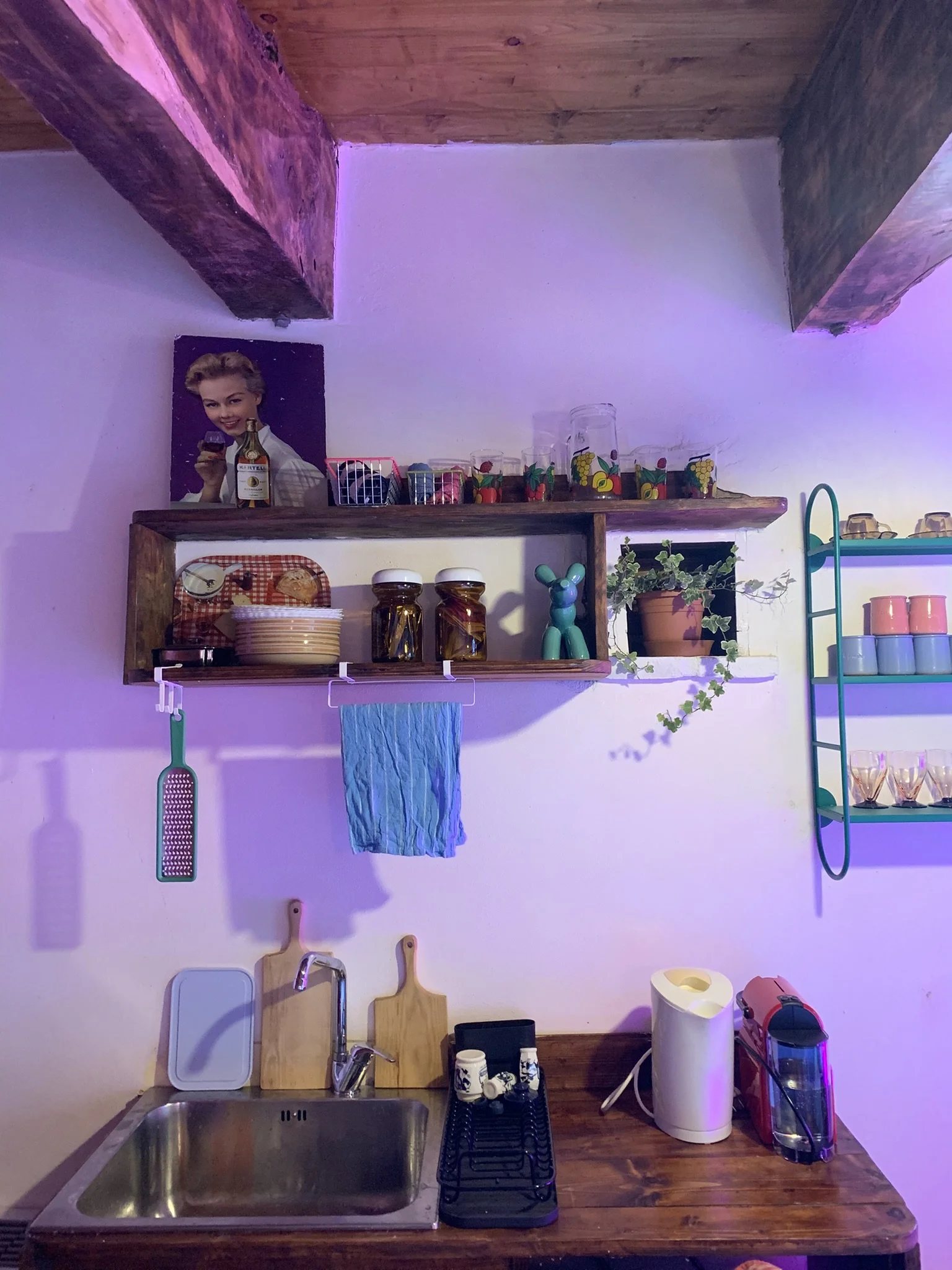 A small kitchen area with a stainless steel sink, wooden countertop, and various kitchen items including a dish drying rack, cups, and kitchen appliances. There are shelves with glassware and decorative items, a towel hanging below the shelves, and a