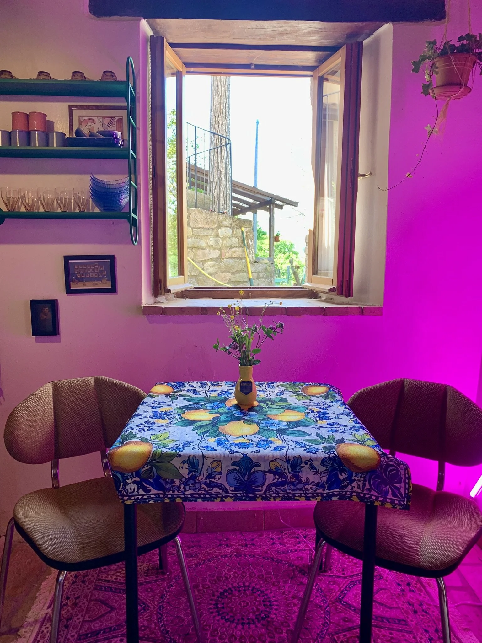 A dining area with a table covered in a colorful tablecloth featuring lemons and blue flowers. Four chairs, two with beige upholstery, surround the table. A yellow vase with greenery is on the table. An open window shows an outdoor stone structure an