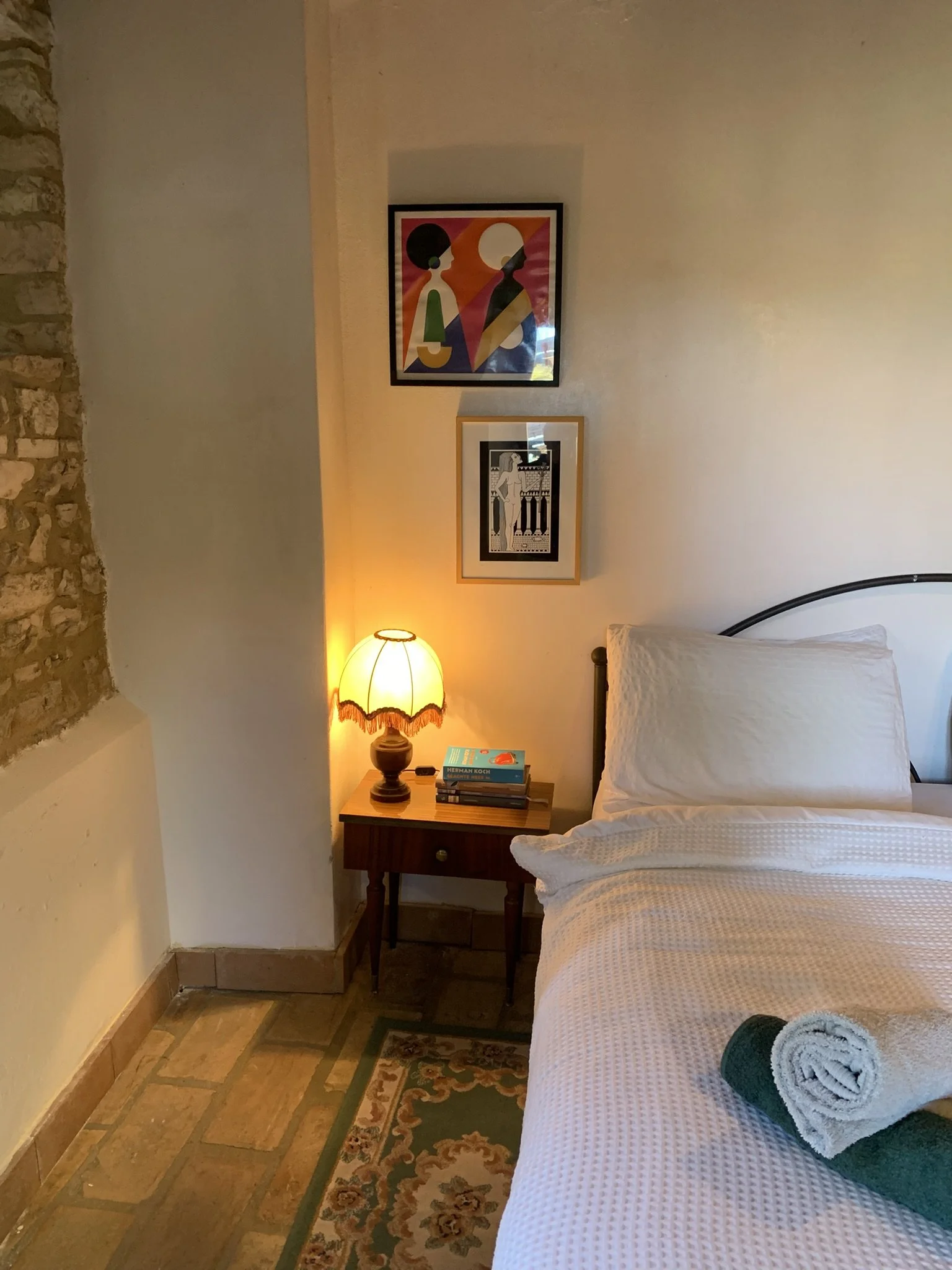 A cozy bedroom with a bed, a nightstand with a lamp, books, and a small framed picture. The room has exposed brick and painted wall, and a rug on a wooden floor. Two framed artworks hang on the wall above the bed.