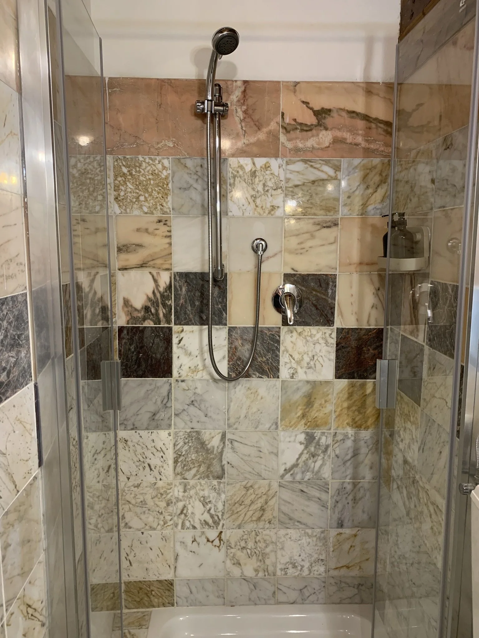 A shower with walls covered in multicolored marble tiles and a glass door. The shower has a handheld showerhead and a soap dispenser on the right side.