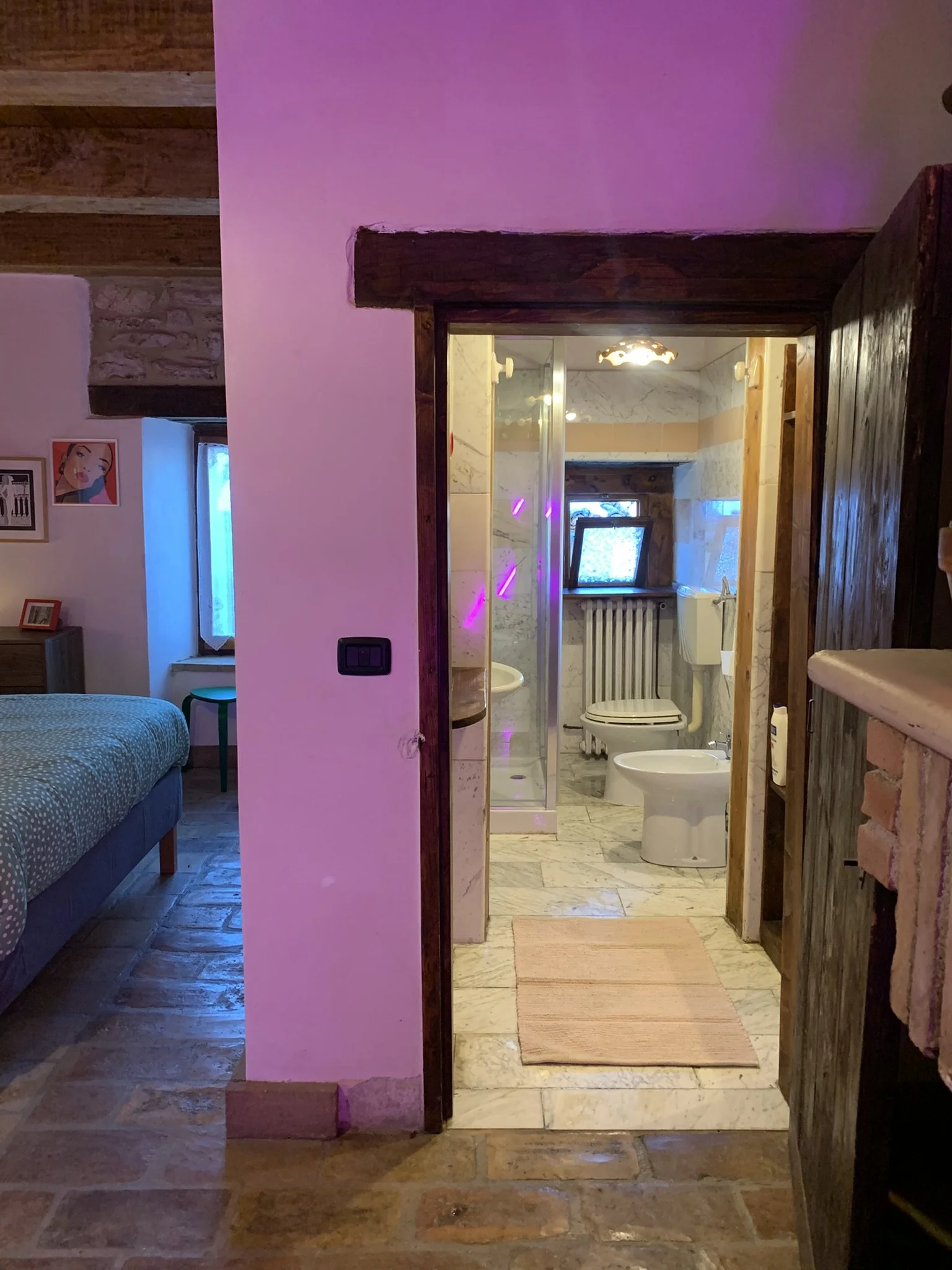 View of a small bathroom with marble floors, a shower, a toilet, and a bidet, seen through an open wooden door from a bedroom with a bed, side table, and artwork on the wall.