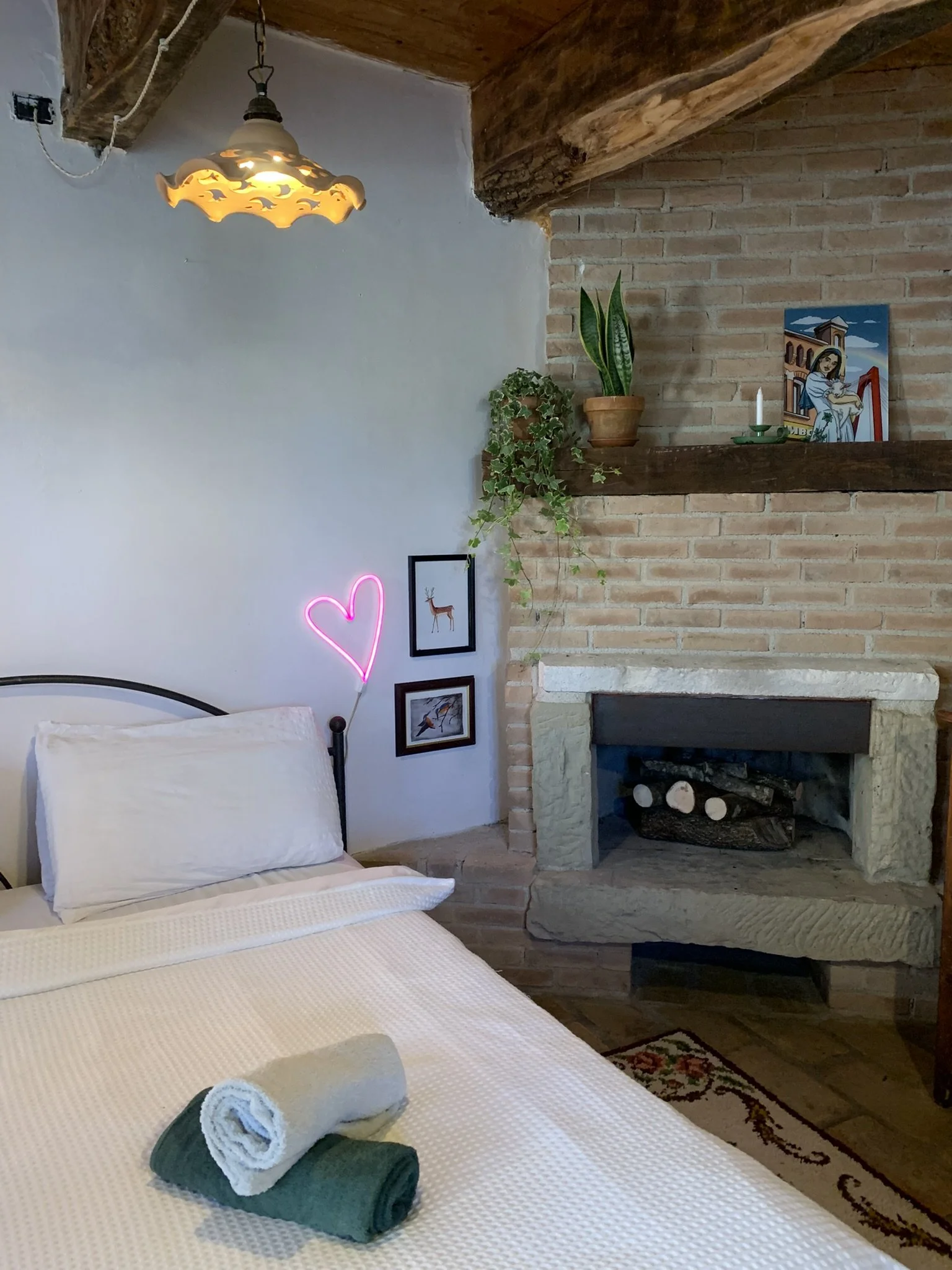 A cozy bedroom with a simple bed, two rolled towels on the bed, a fireplace with a brick and wood mantel, a potted plant, a candle, a small painting, and a neon pink heart light on the wall.