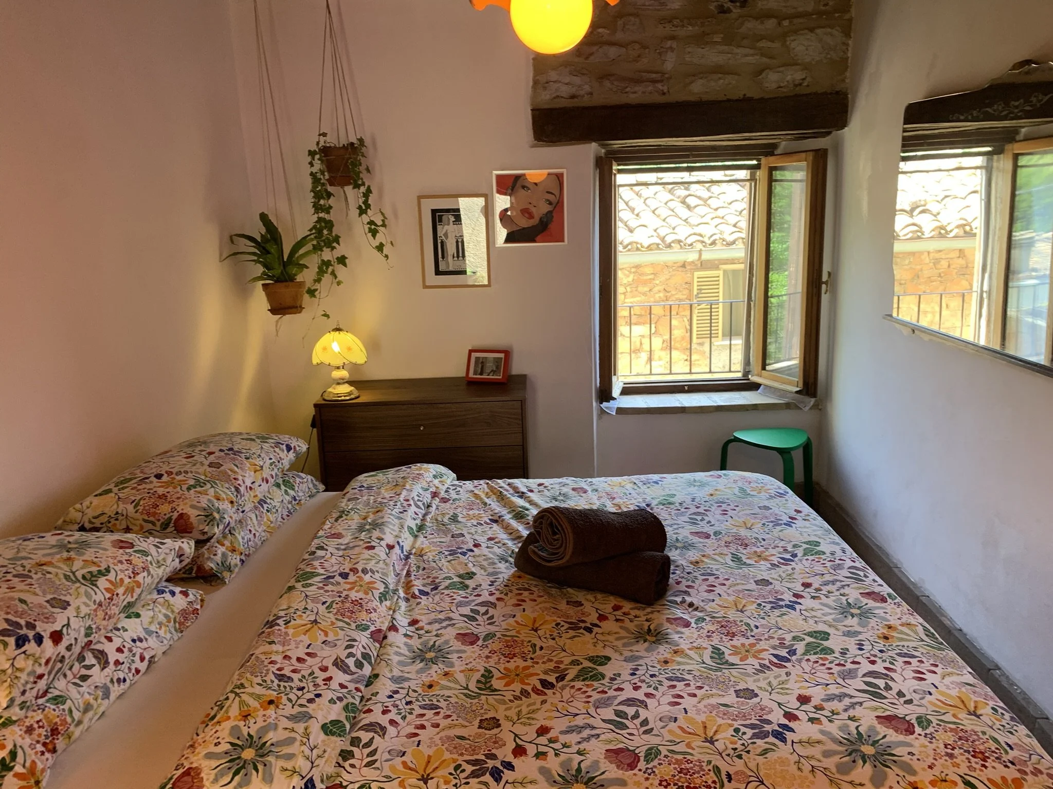 A bedroom with two beds covered in floral-patterned bedsheets. There are neatly rolled brown towels on one bed. The room features a wooden desk with a small yellow lamp and framed pictures on its surface. An open window with wooden frames shows a vie