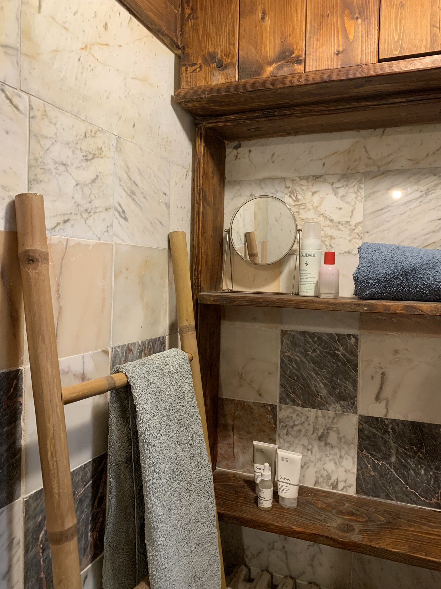A bathroom corner with a wooden shelf holding toiletries, a small round mirror, a blue towel, a wooden ladder with a gray towel, and tiled walls with varied marble patterns.
