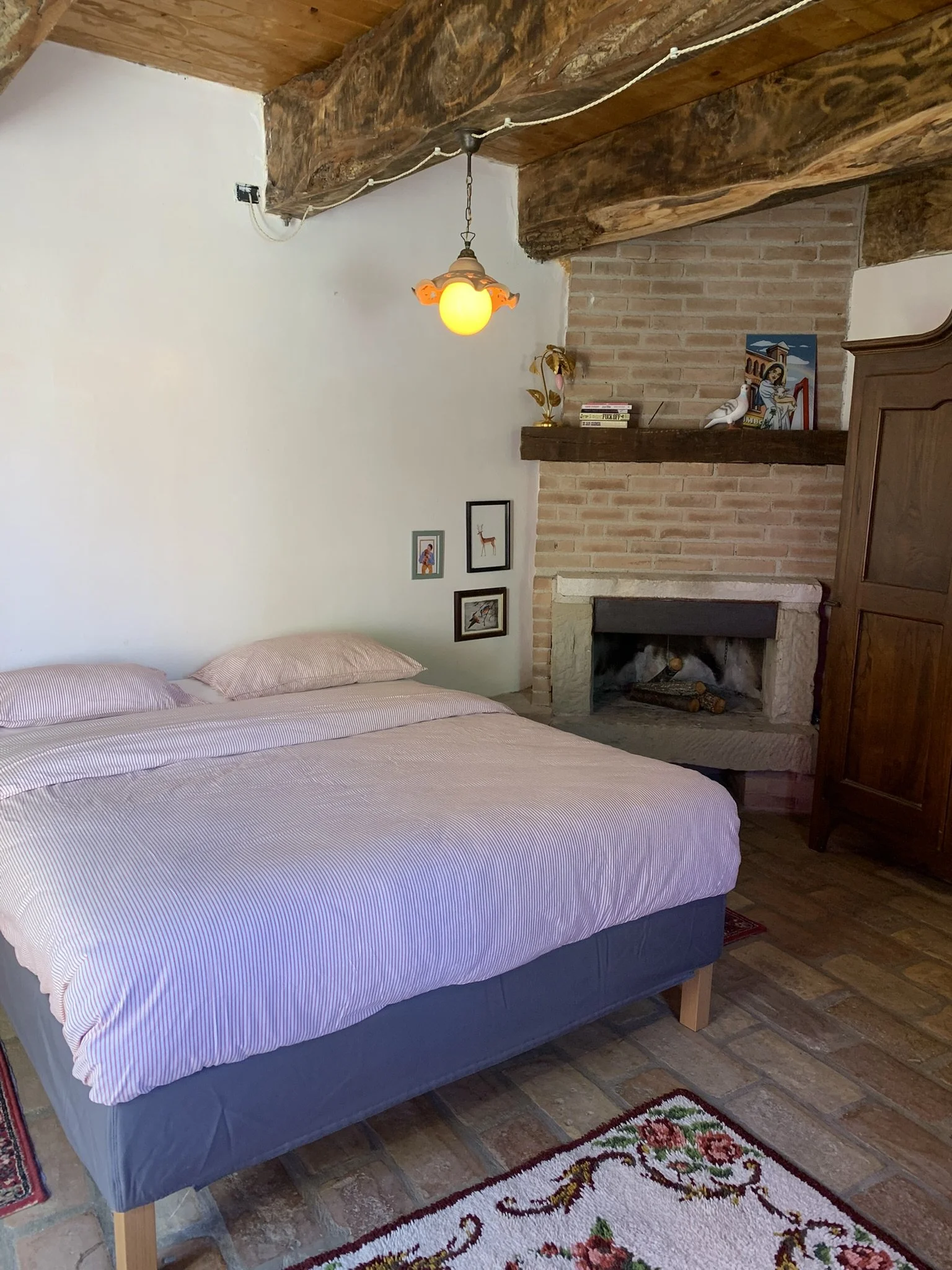A bedroom with a bed that has a pink and white striped cover and pillows, a brick fireplace with logs, a wooden mantel with decorative items, and a small colorful rug on a wooden floor.