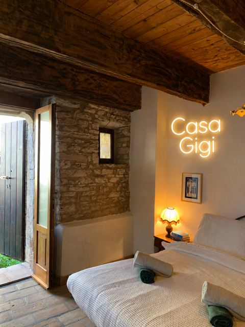 Cozy bedroom with stone wall, wooden ceiling beams, and a neon sign that reads 'Casa Gigi'. There is a bed with rolled towels, a small nightstand with a lamp, and a doorway leading outside.