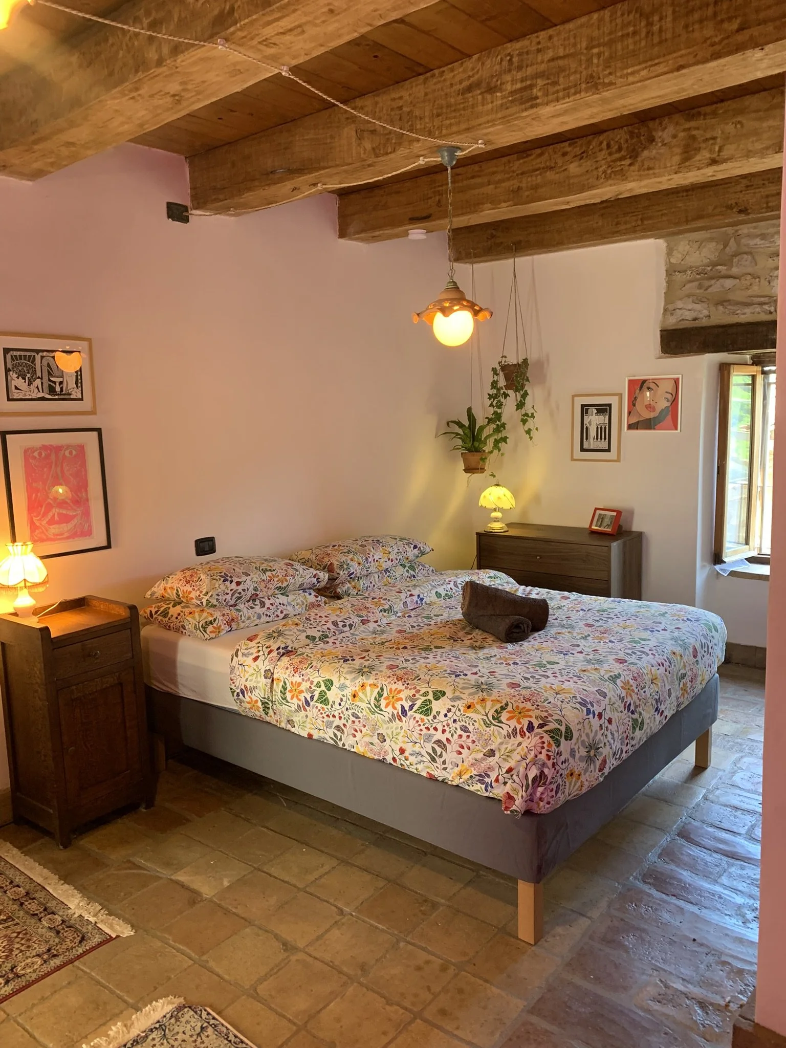 A cozy bedroom with a wooden beam ceiling, a double bed with floral bedding, a nightstand with a small lamp, and wall art. A window is open, letting in natural light.