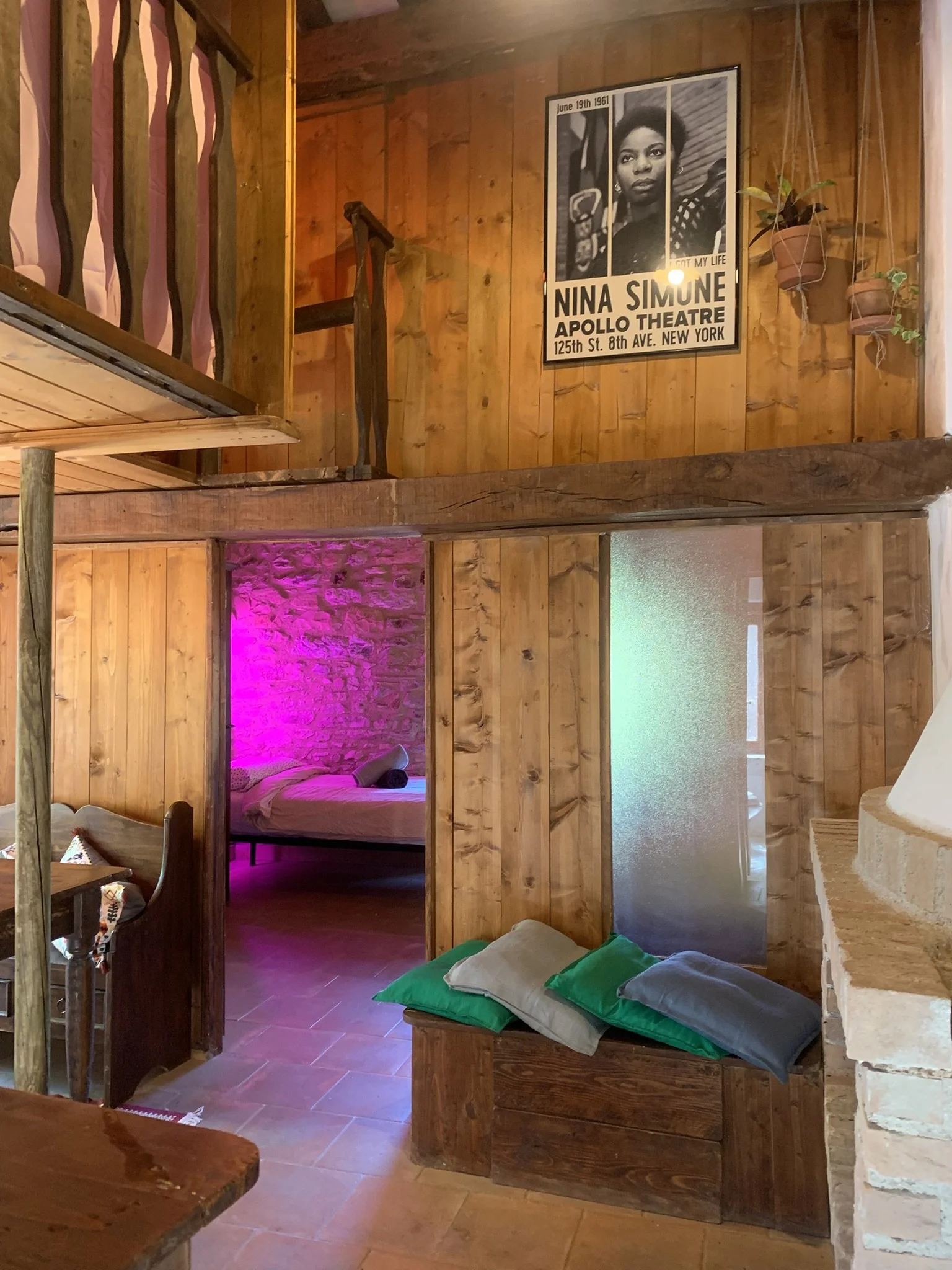 Interior of a wooden room with a poster of Nina Simone on the wall, some hanging potted plants, a bed with a rolled towel, and a staircase going up.