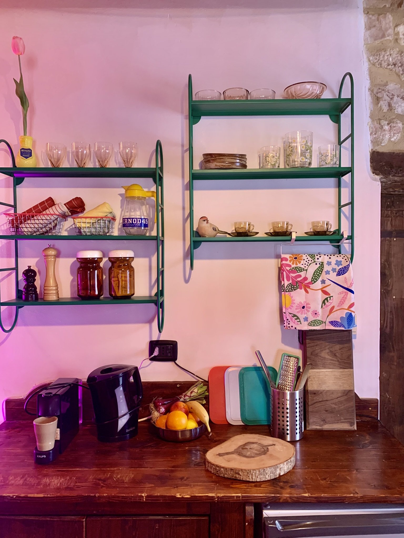 Kitchen counter with coffee maker, fruit bowl with oranges and bananas, various kitchen utensils, and shelves with glassware and dishes.