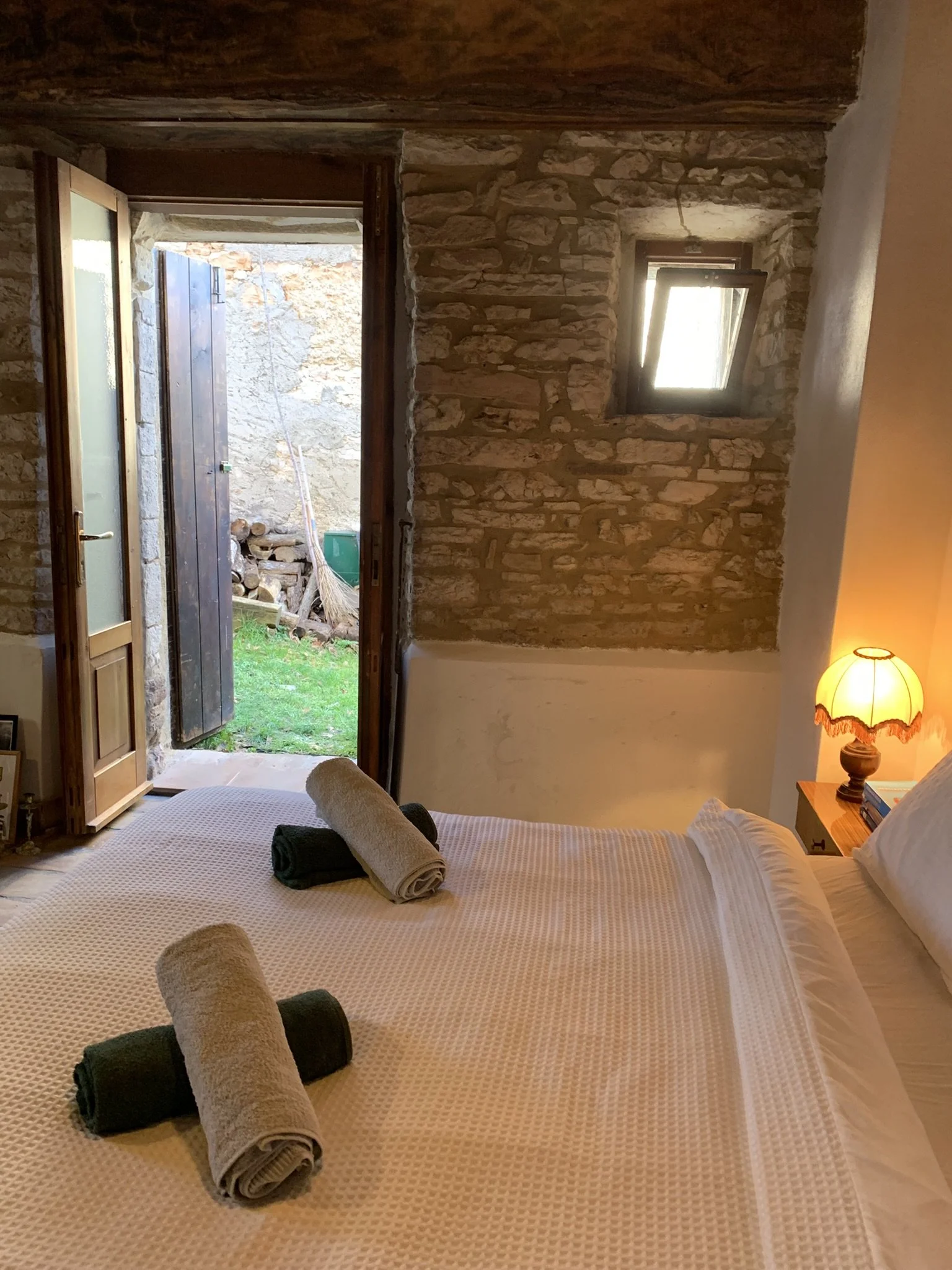 A cozy bedroom with a bed covered in a textured beige bedspread, two rolled towels on top, and a nightstand with a lamp. An open door reveals an outdoor yard area with stacked firewood, a broom, and a green bucket. The walls are made of rustic stone,
