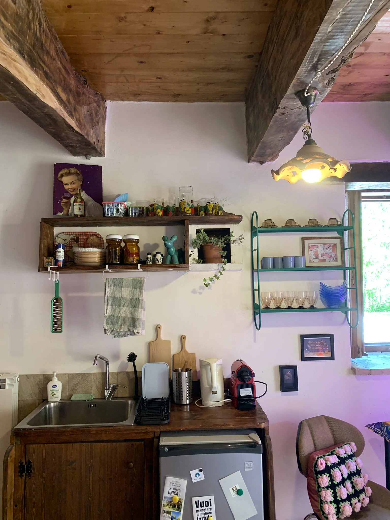 A kitchen with wooden shelves, a small refrigerator, a sink, various kitchen utensils, and a chair with a pink floral blanket. The wall has a photo of a woman holding a glass of red wine and a bottle, along with some framed pictures and colorful cups