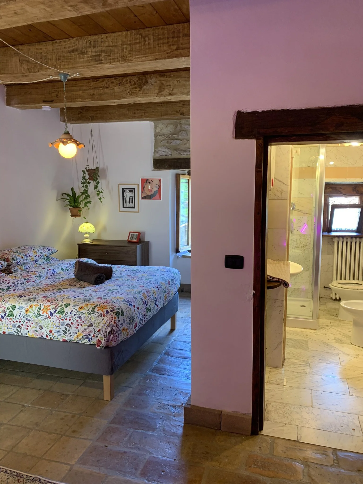 A bedroom with a bed that has a colorful floral bedspread, and a towel rolled on top. There are framed pictures on the wall, hanging plants, a bedside lamp, and a wooden dresser. To the right is an opening to a bathroom with a shower stall, toilet, a