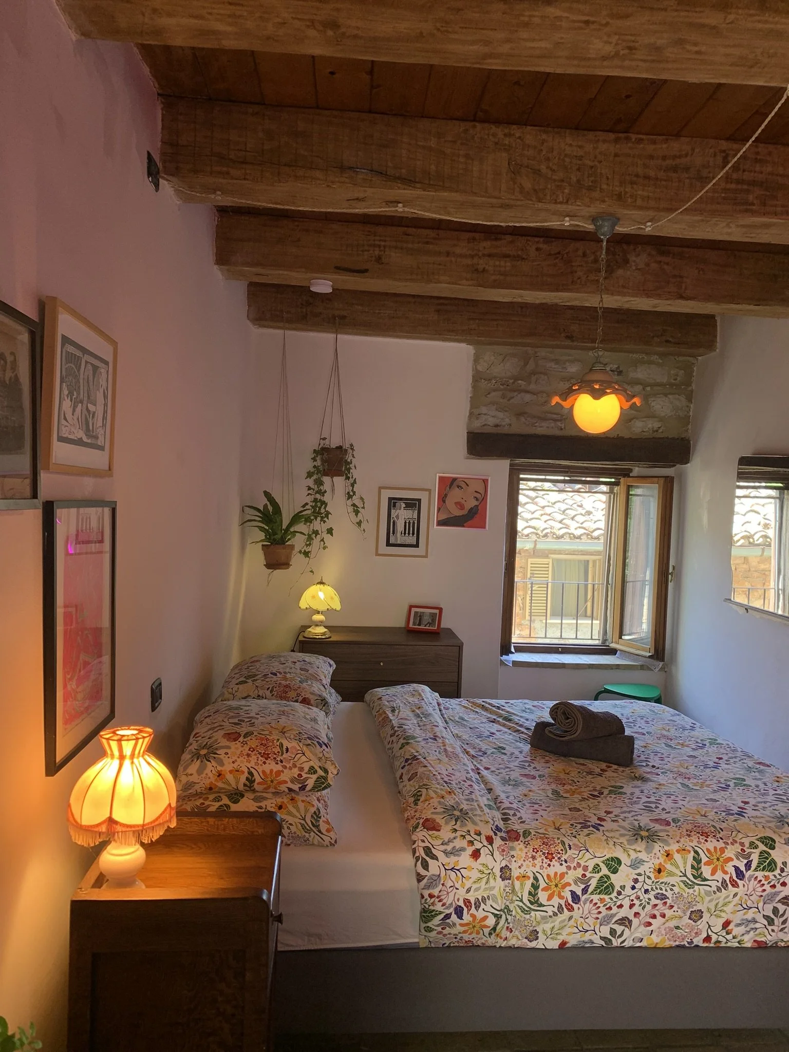 A cozy bedroom with a wooden ceiling and exposed beams. There's a large window with open shutters letting in natural light. The bed has floral patterned bedding with rolled towels on top. A nightstand with a lit table lamp is beside the bed, and vari
