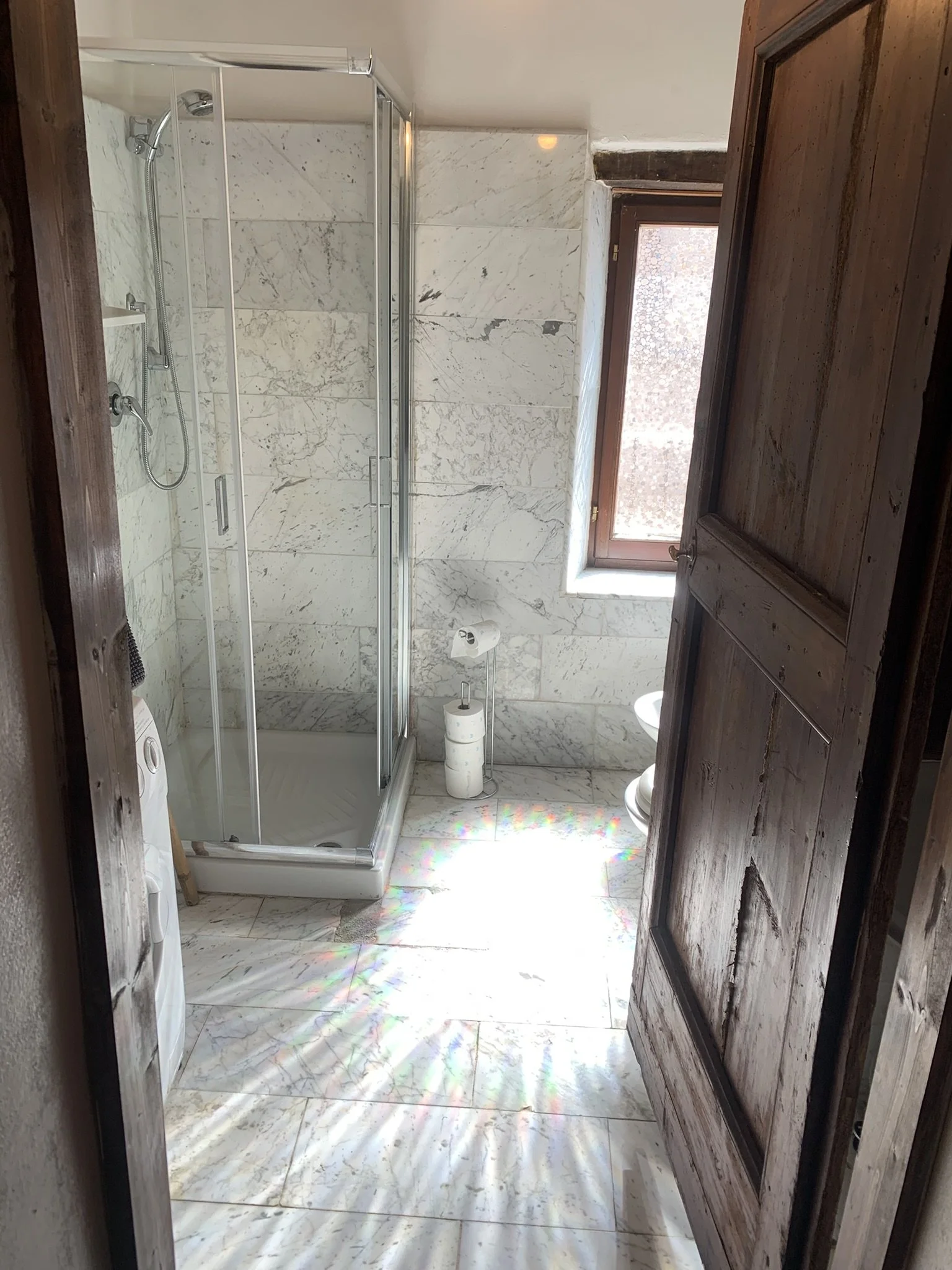Small bathroom with marble floor and walls, wooden door open to reveal a shower with glass door, window with natural light, and a toilet with toilet paper rolls.