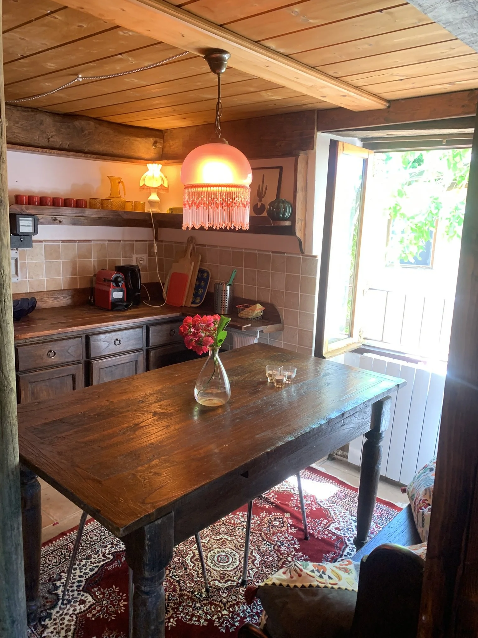 A rustic kitchen scene with a dark wooden table in the foreground, a red floral centerpiece, and traditional patterned rugs underneath. There's a wooden cabinet on the left and a shelf with decorative items such as a lamp, vases, and candles on the w