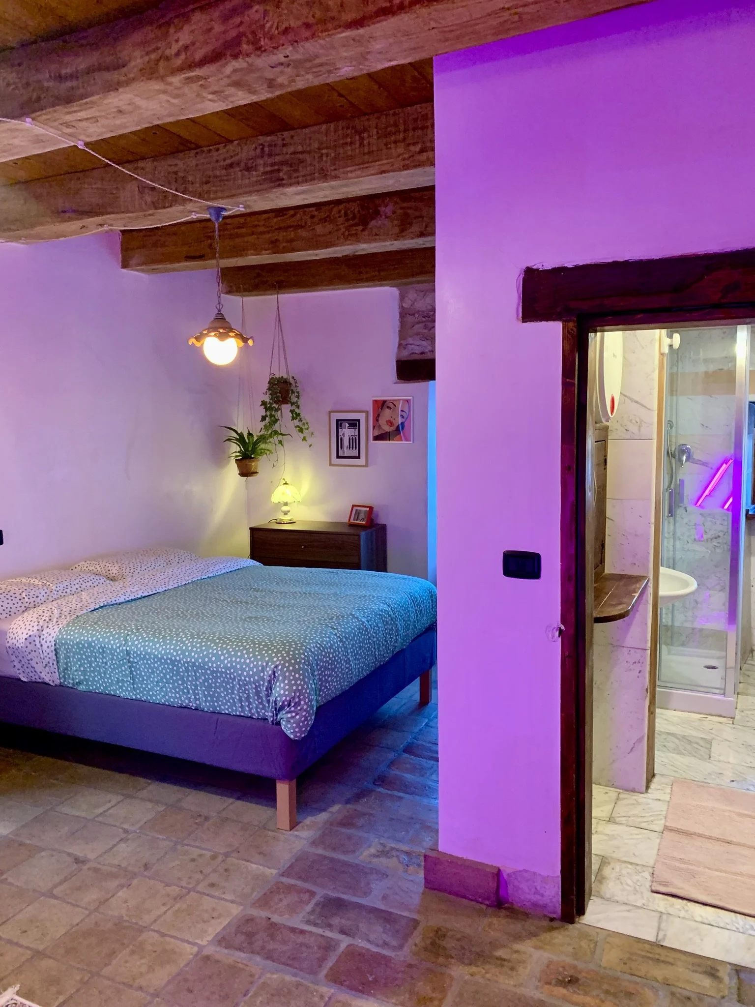 A bedroom with a bed, nightstand, and lamps, with an adjacent bathroom visible through an open doorway.
