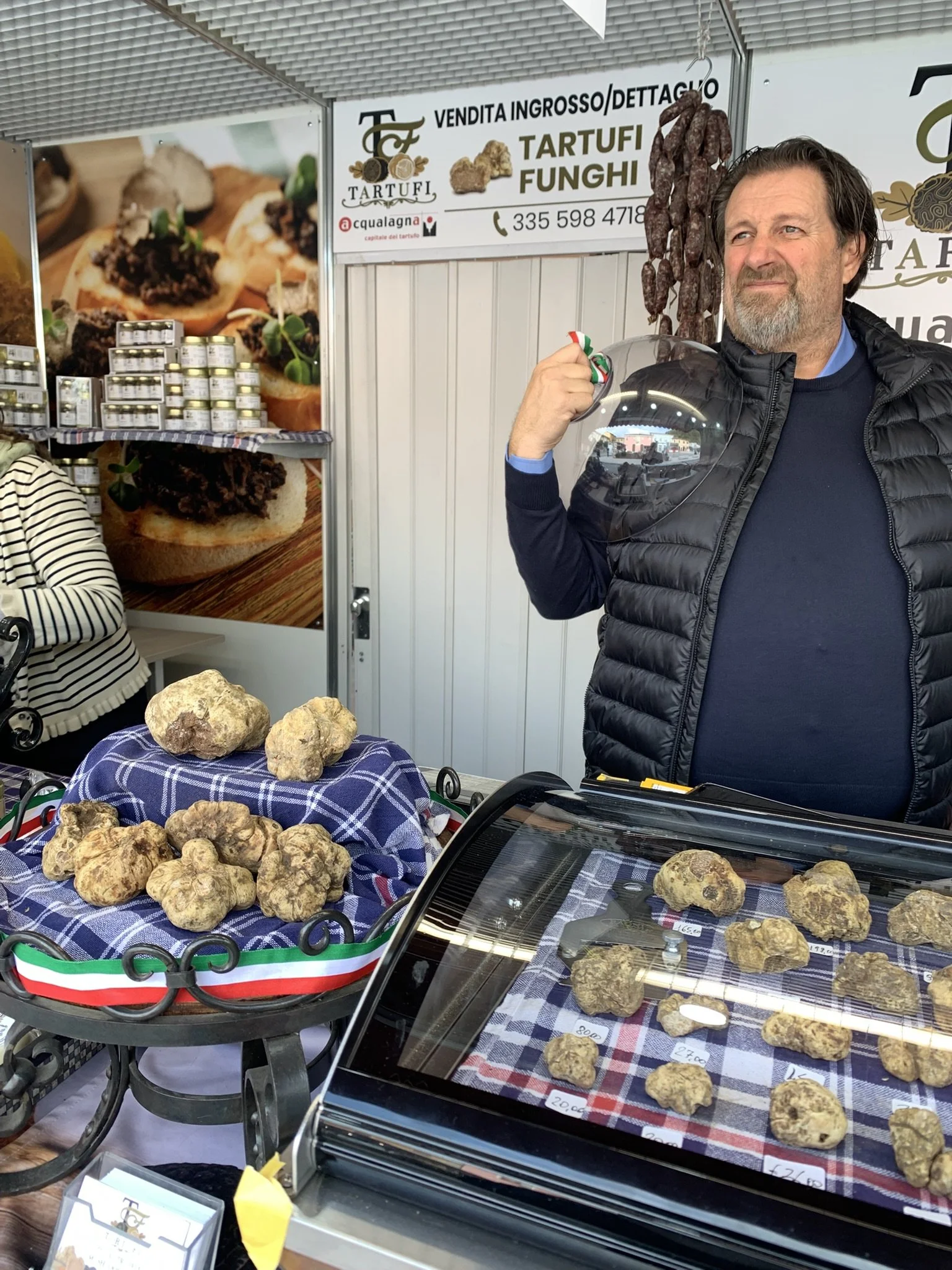A man holding a mushroom at an outdoor market stall with dried sausages, jars of truffle products, and truffles on display.