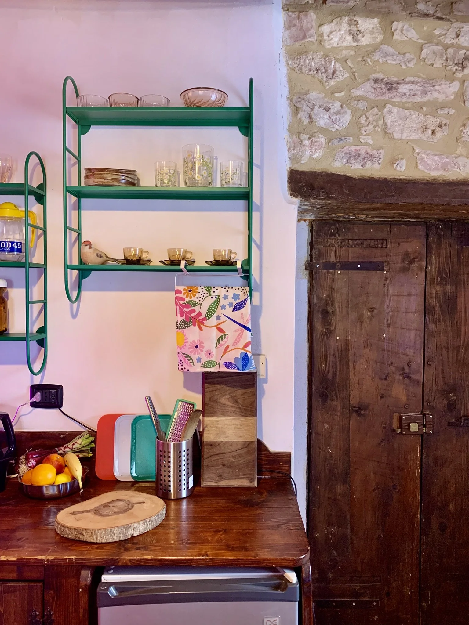 Kitchen shelf with glassware, plates, and cups, placed next to a stone wall and a wooden door. There are colorful cutting boards, a fruit bowl, and kitchen utensils on the wooden countertop below.
