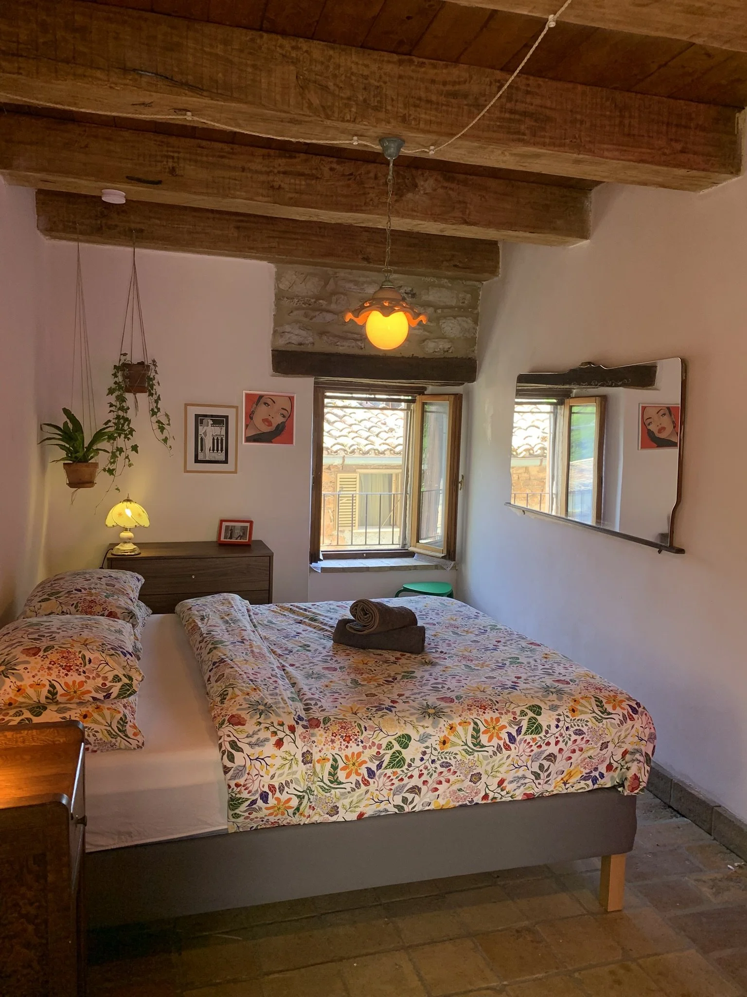 A cozy bedroom with a large bed covered in a colorful floral duvet, two rolled towels on top, and matching floral pillows. There is a wooden bedside table with a small yellow lamp, and a potted plant hanging from the ceiling. The room features wooden