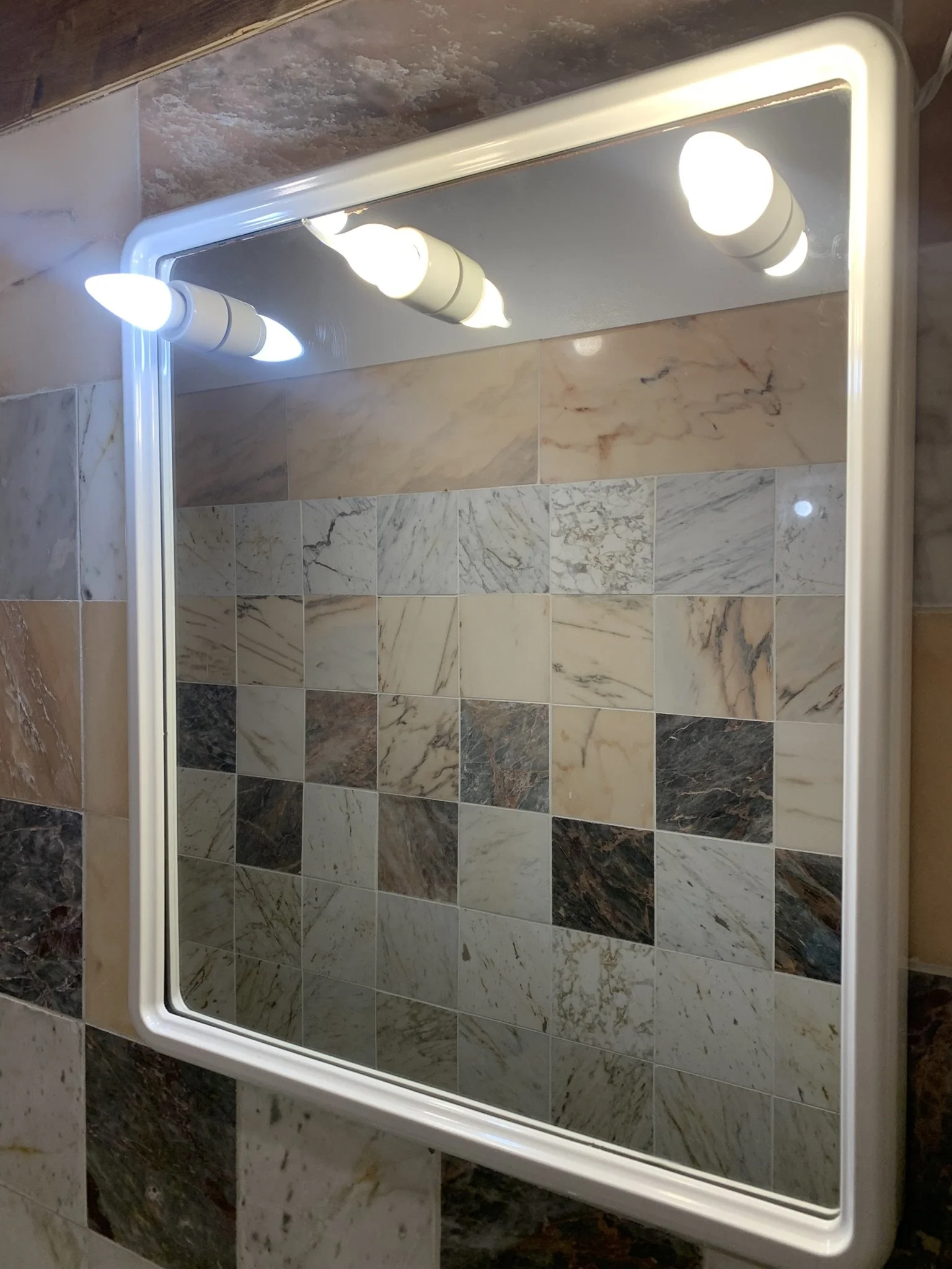 A rectangular mirror with a white frame mounted on a wall. Reflected in the mirror is a tiled wall with a pattern of variously colored marble-like tiles and a ceiling light fixture with three frosted glass bulbs.