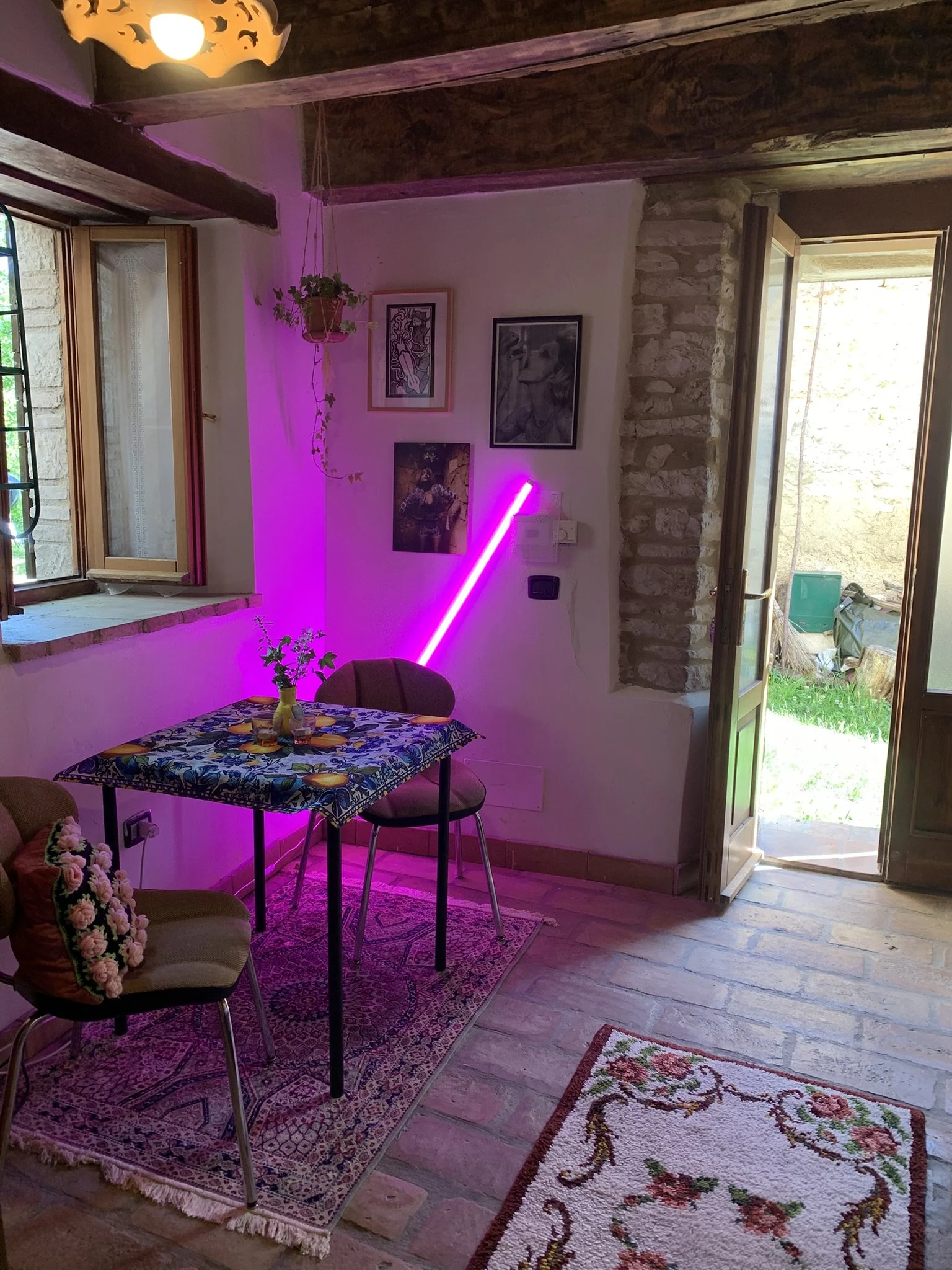 A cozy room with a small table covered with a vibrant, patterned tablecloth, and two chairs. There are several framed pictures on the wall, a hanging plant, a pink neon light, and an open door revealing a garden outside. The room has a wooden ceiling