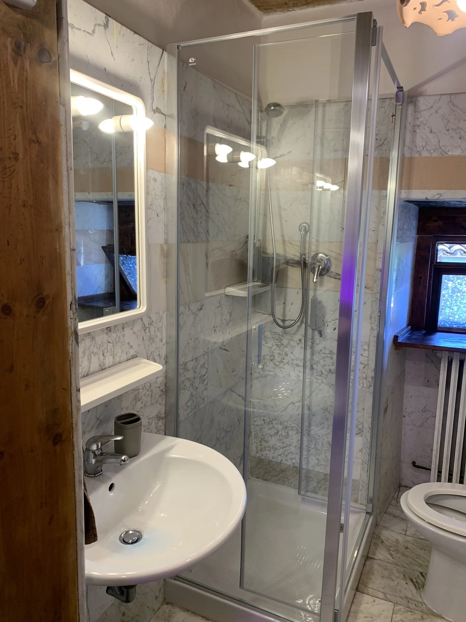 A small bathroom with marble walls, a mirror with lights, a corner shower enclosure, a white sink with a faucet and a small trash bin, and a toilet. There is a wooden wall on the left and a window with a blue patterned curtain on the right.
