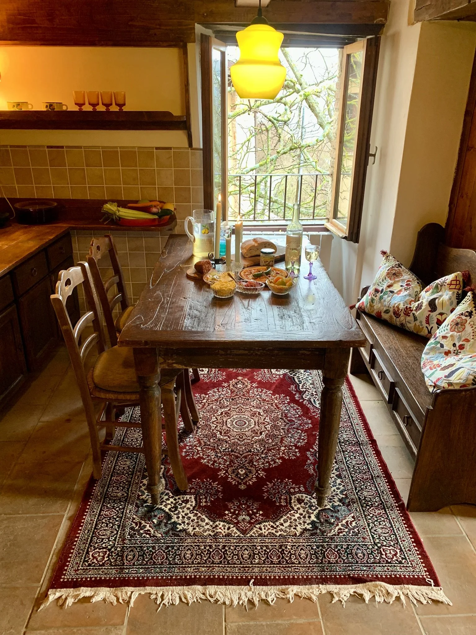 A dining room with a wooden table, six chairs, a red patterned rug, and a bench with a colorful pillow. The table is set with food, drinks, and candles. There is a large window open to the outdoors with green trees visible. The room has wooden accent