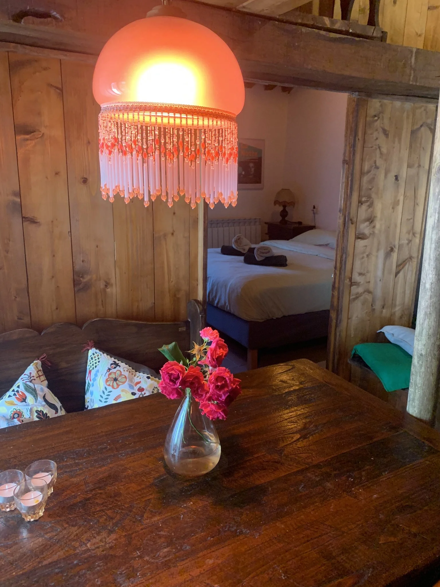 A cozy bedroom with wooden walls, a bed with rolled towels, a wooden nightstand with a lamp, and a large red and pink beaded chandelier hanging from the ceiling. There is a vase with pink flowers on a wooden table in the foreground.