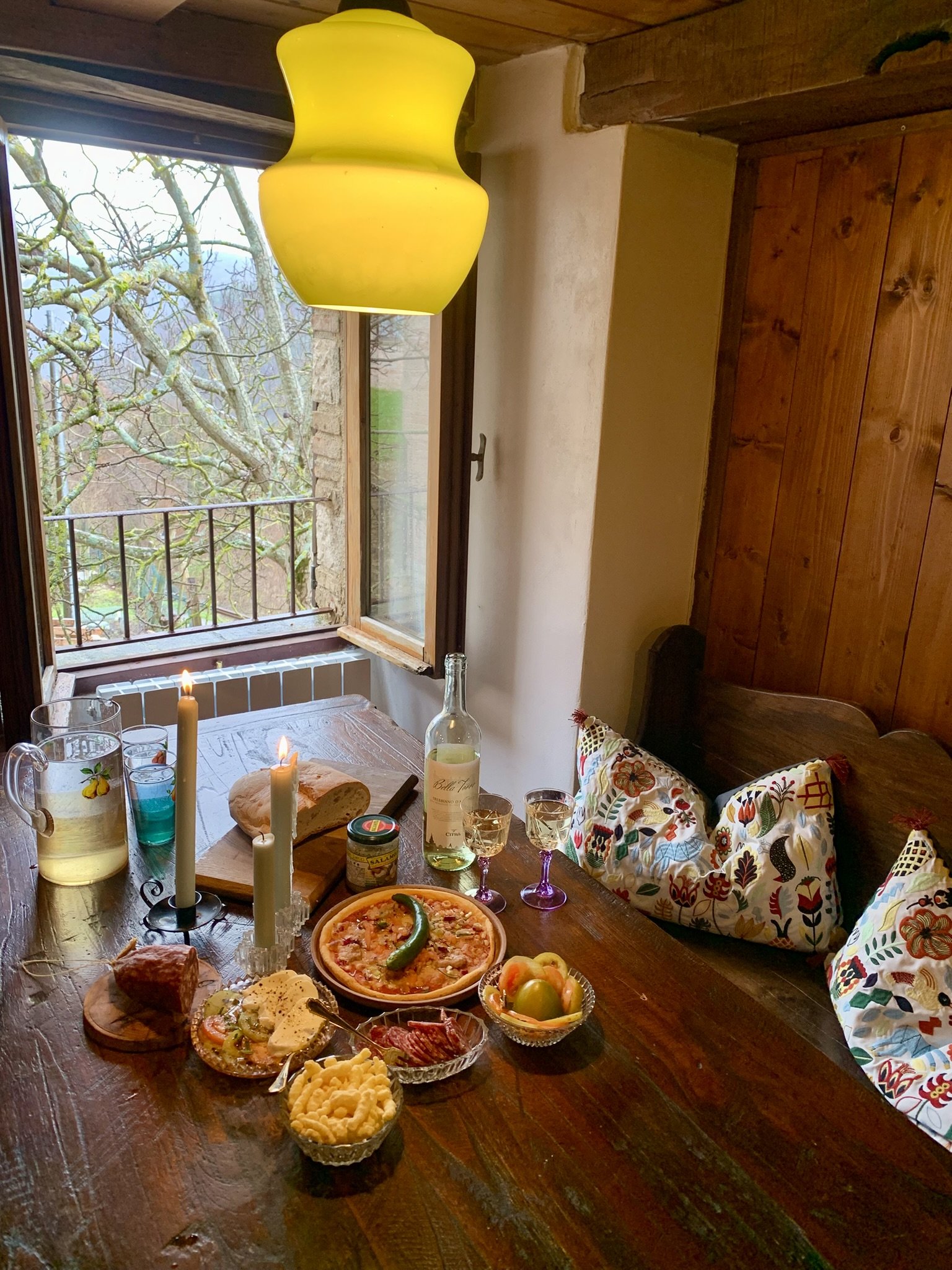 A cozy dining area with a wooden table set for a meal, featuring a pizza, various appetizers, drinks, and lit candles. An open window reveals a view of a tree with bare branches outside, and colorful cushions are on a bench along the wall.