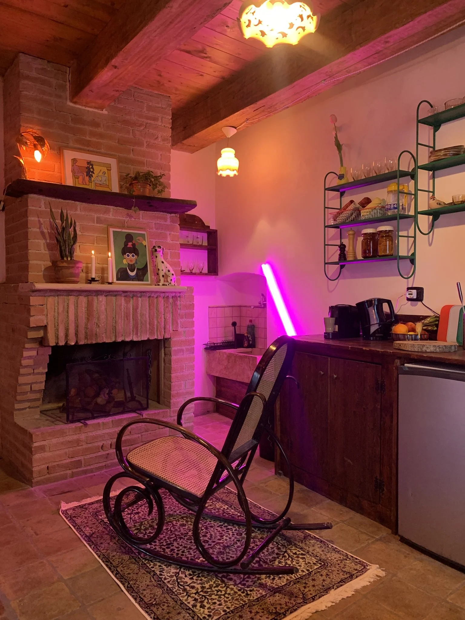 A cozy kitchen area with a brick fireplace, wooden ceiling, and warm lighting. There is a small rectangular rug with intricate patterns on the floor, surrounded by a black rocking chair. The brick fireplace has decorative plants and framed artwork on