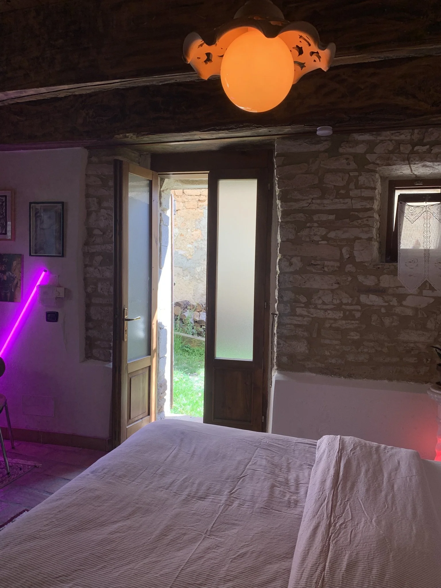 A bedroom with a bed covered in beige striped bedding, stone walls, an open door leading outside with sunlight, a ceiling lamp, and purple and orange neon lighting.
