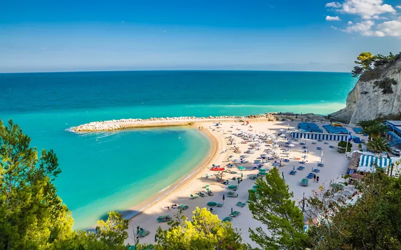 Aerial view of a crescent-shaped beach with turquoise water, white sand, and a stone jetty, surrounded by green trees and a small resort area with striped tents and umbrellas.