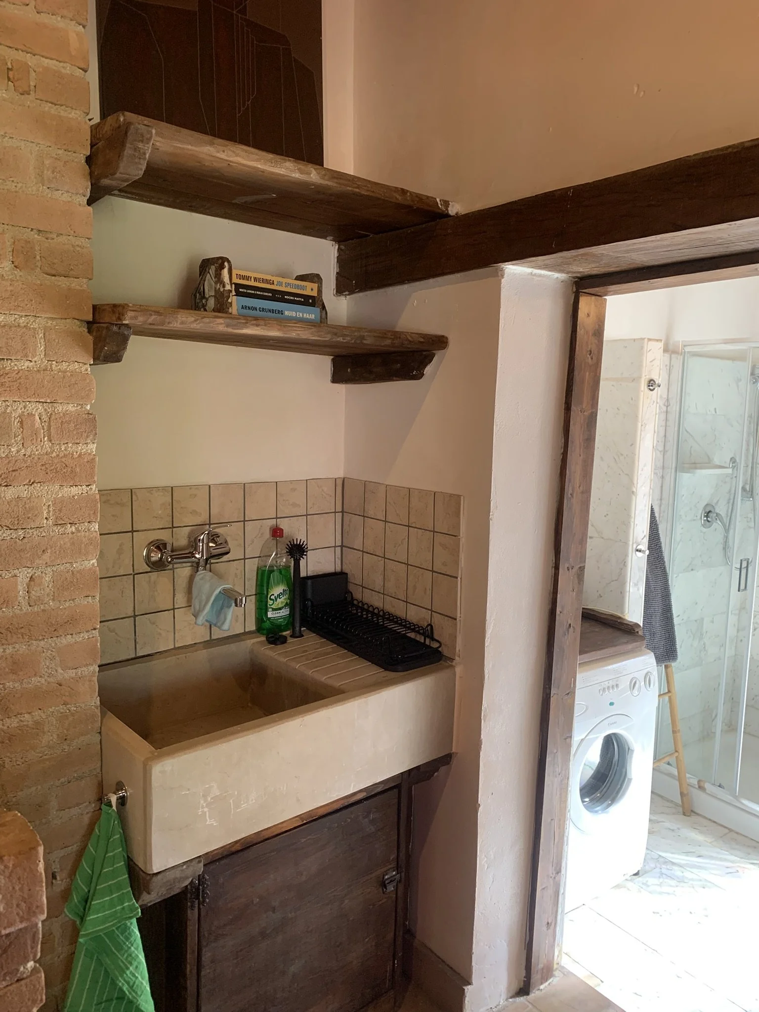 A small bathroom area with a rustic wooden countertop and a deep, stone-like basin sink. A silver faucet is mounted above the sink. To the right is a view into a laundry area with a washing machine, a wooden stool, and a walk-in shower with glass doo