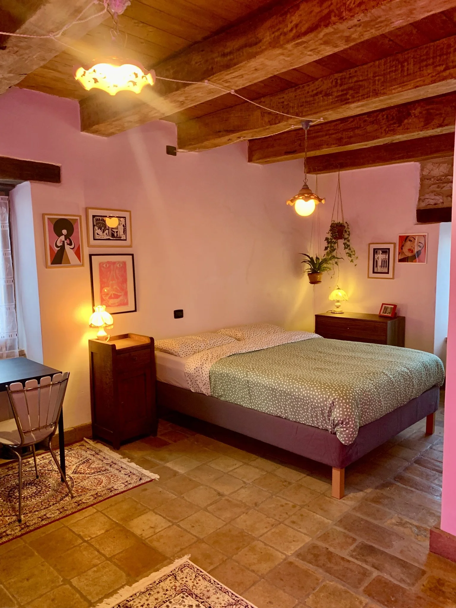 A cozy bedroom with a bed covered in a polka-dot green cover and white sheets with black dots, two bedside lamps emitting warm light, artwork on the pink walls, hanging plants, wood-beamed ceiling, and tile floor.