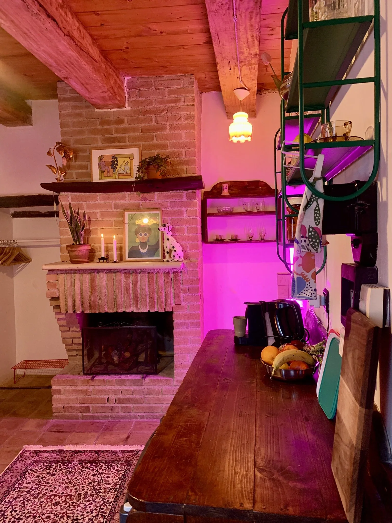 A cozy kitchen with a brick fireplace, decorated with framed artwork and potted plants. A wooden countertop holds a bowl of fruit, a to-go coffee cup, and some kitchen utensils. Shelves with glasses and dishes are illuminated with pink, purple, and w