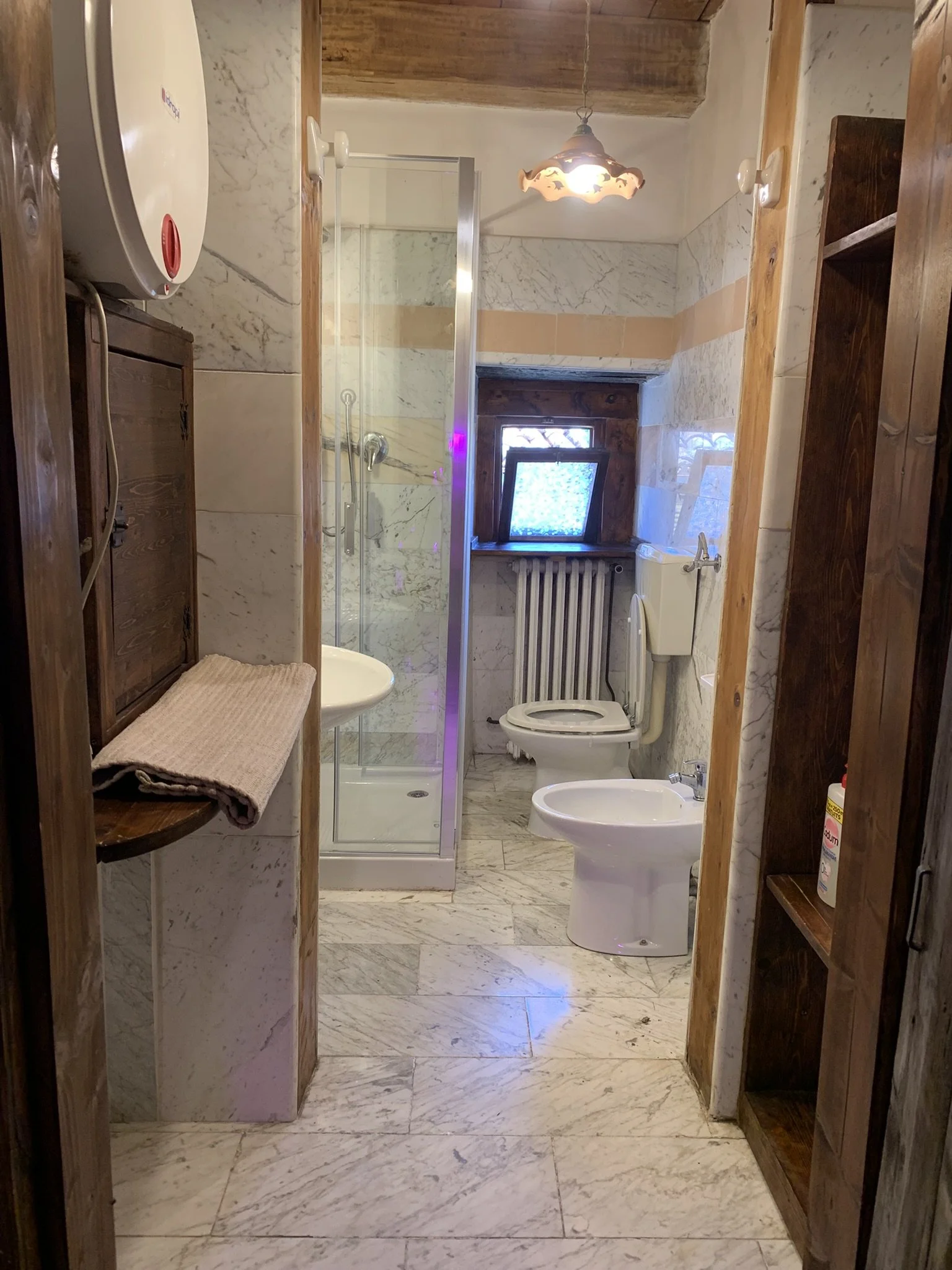 A small bathroom with marble walls and floors, a shower stall, a toilet, a bidet, a small window, and wooden shelves and cabinets.