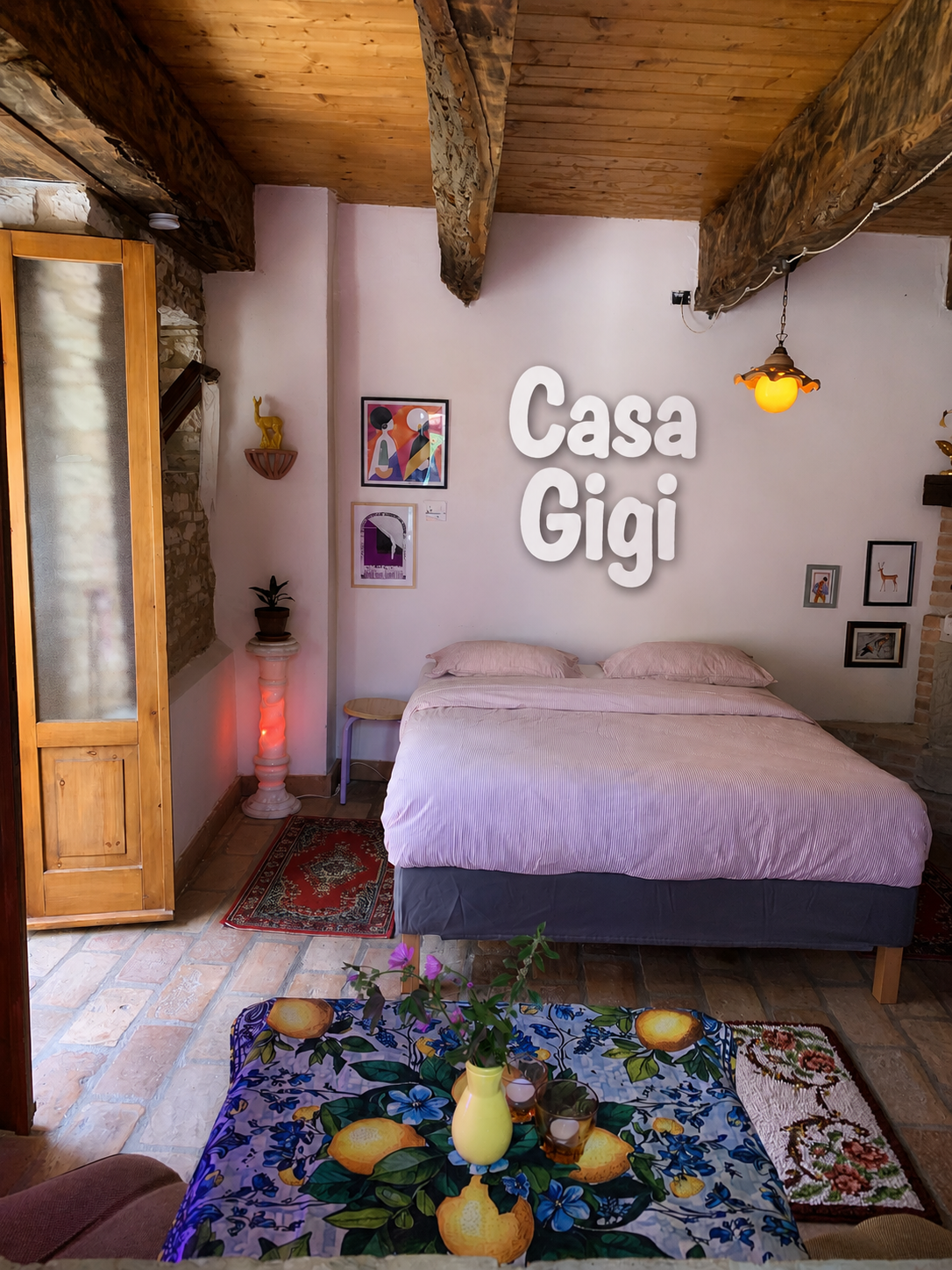 A cozy bedroom with a bed covered in light pink sheets and pillows, a colorful table with a lemon and flower design, a small stool, framed artwork on the walls, and a wooden ceiling with exposed beams. The words 'Casa Gigi' are displayed on the wall.