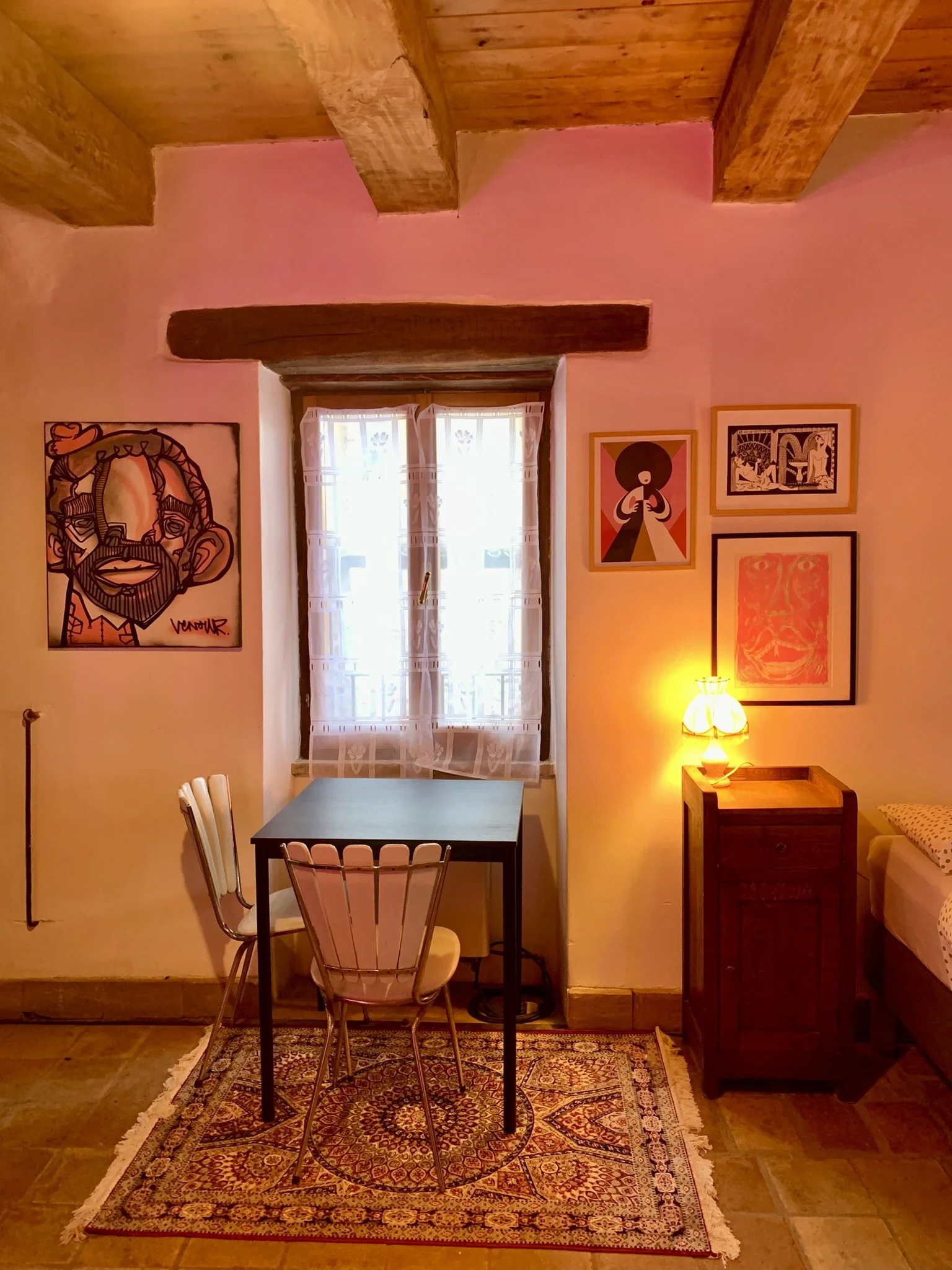 Interior of a cozy room with pink walls, wooden beam ceiling, and a window with sheer curtains. The room features artwork on the walls, a small table with white chairs, a rug, a side table with a lamp, and part of a sofa with a polka dot pillow.