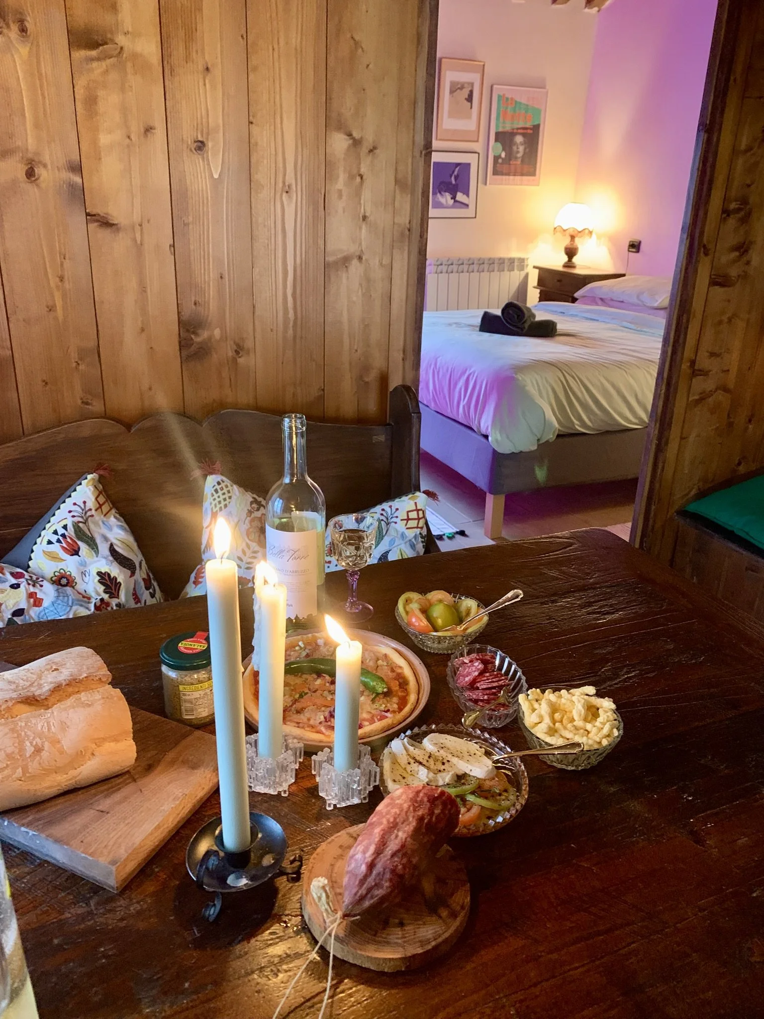 A wooden dining table set with various foods including a pizza, bread, cured meats, cheese, and a bowl of fruit, with candles, a bottle of wine, and glasses. In the background, a bedroom with a bed, nightstand, lamp, and framed pictures on the wall. 