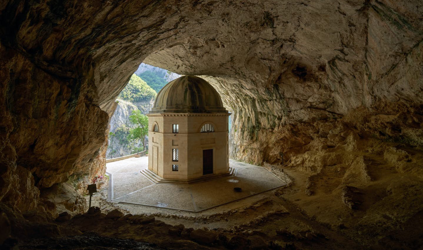 A small building with a domed roof inside a large cave with an opening to the outside, showing trees and mountains.