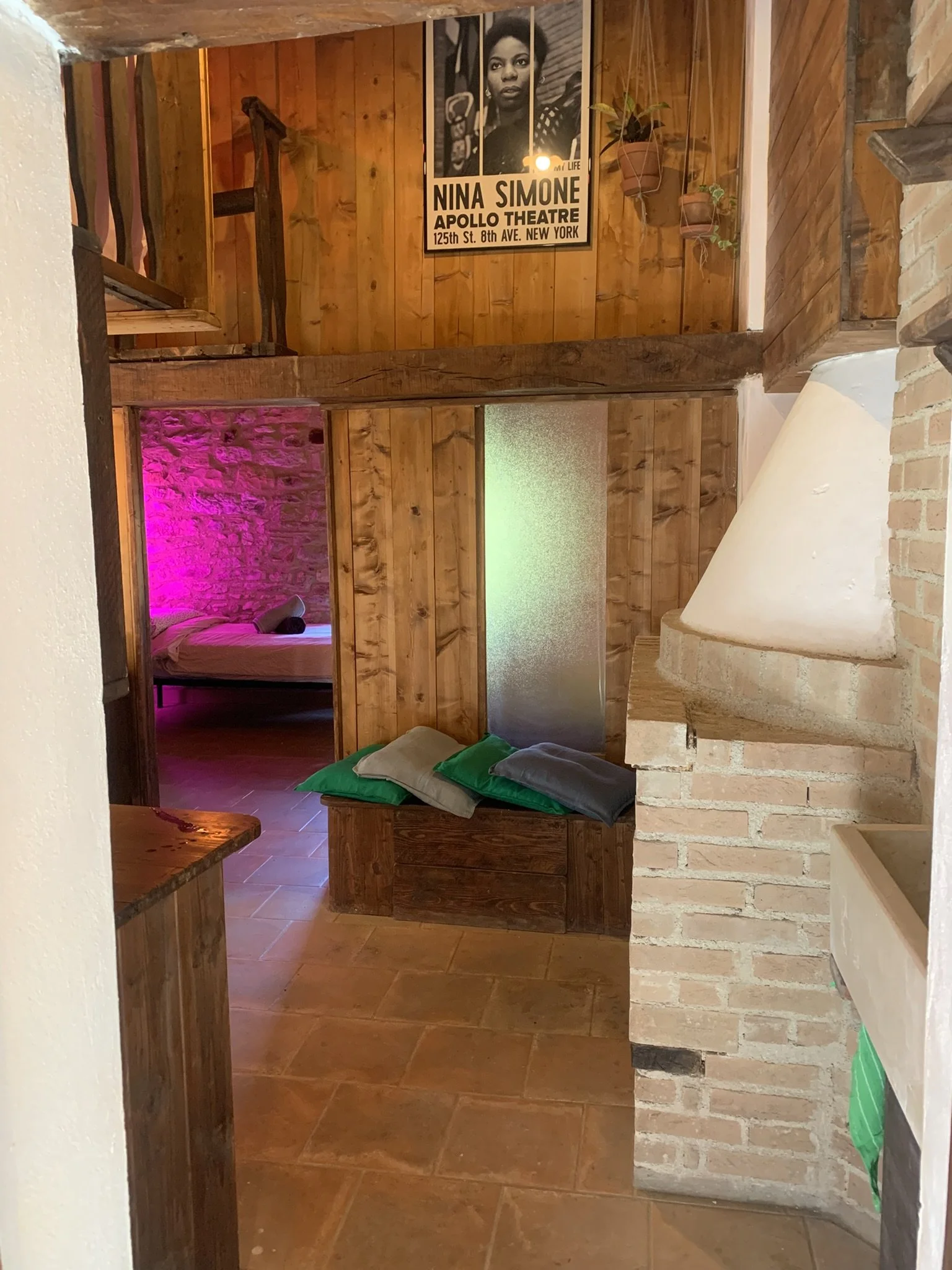A cozy wooden interior with a small bedroom visible through a doorway. The bedroom has a bed with green pillows, pink ambient lighting, and textured stone walls. The main room features wood-paneled walls, hanging potted plants, and a framed black and