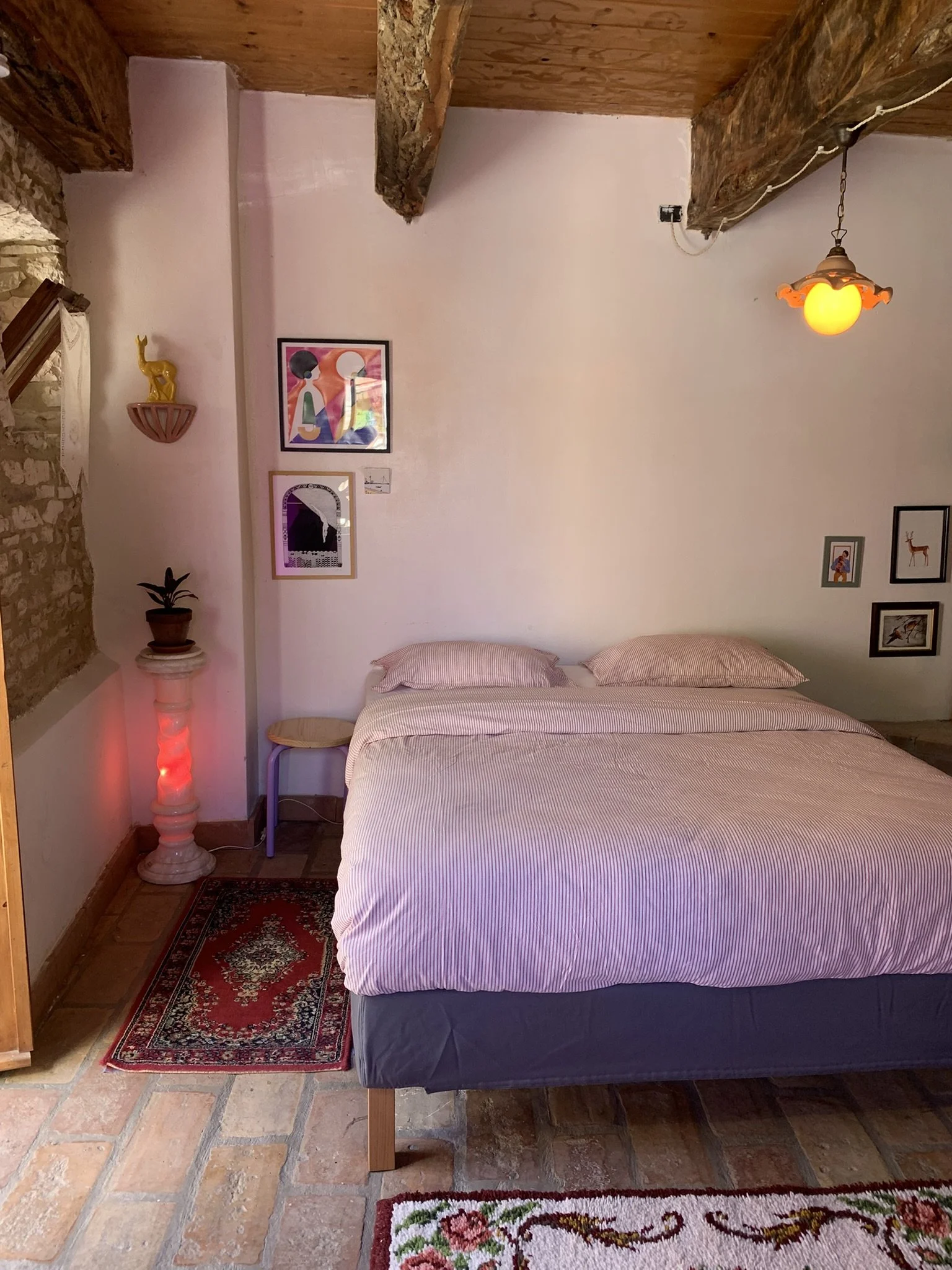 A bedroom with a bed covered in a striped sheet, a small bedside table, a framed picture of a woman, and a small framed silhouette picture on the wall. The room features exposed brick and wooden beams, a hanging light fixture, a potted plant, a decor