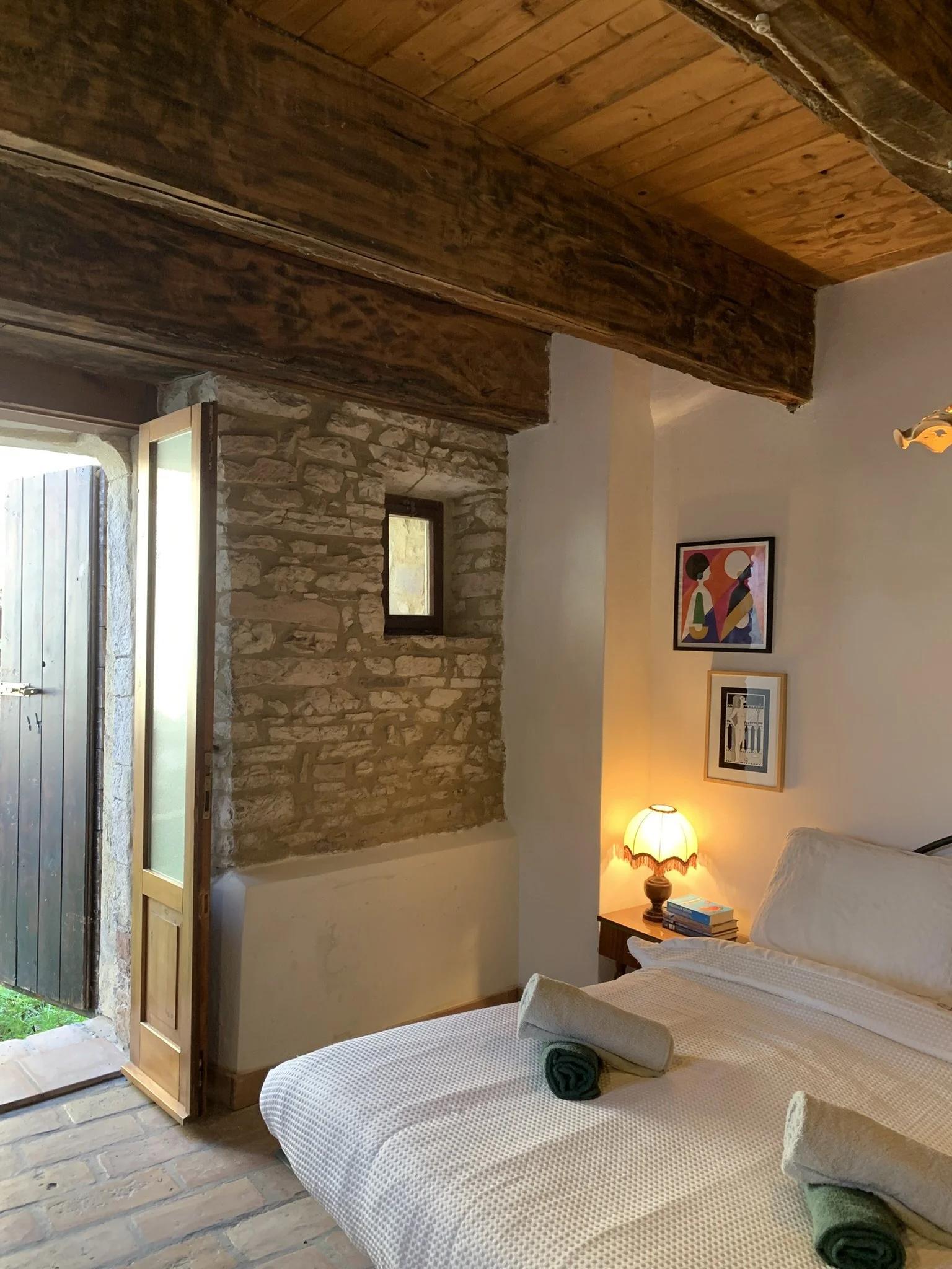 A cozy bedroom with a bed covered in a white knitted bedspread, set against a stone wall with artwork. A small nightstand holding a lamp with a lampshade and stacked books. An open door with a view outside, and an exposed wooden beam ceiling.
